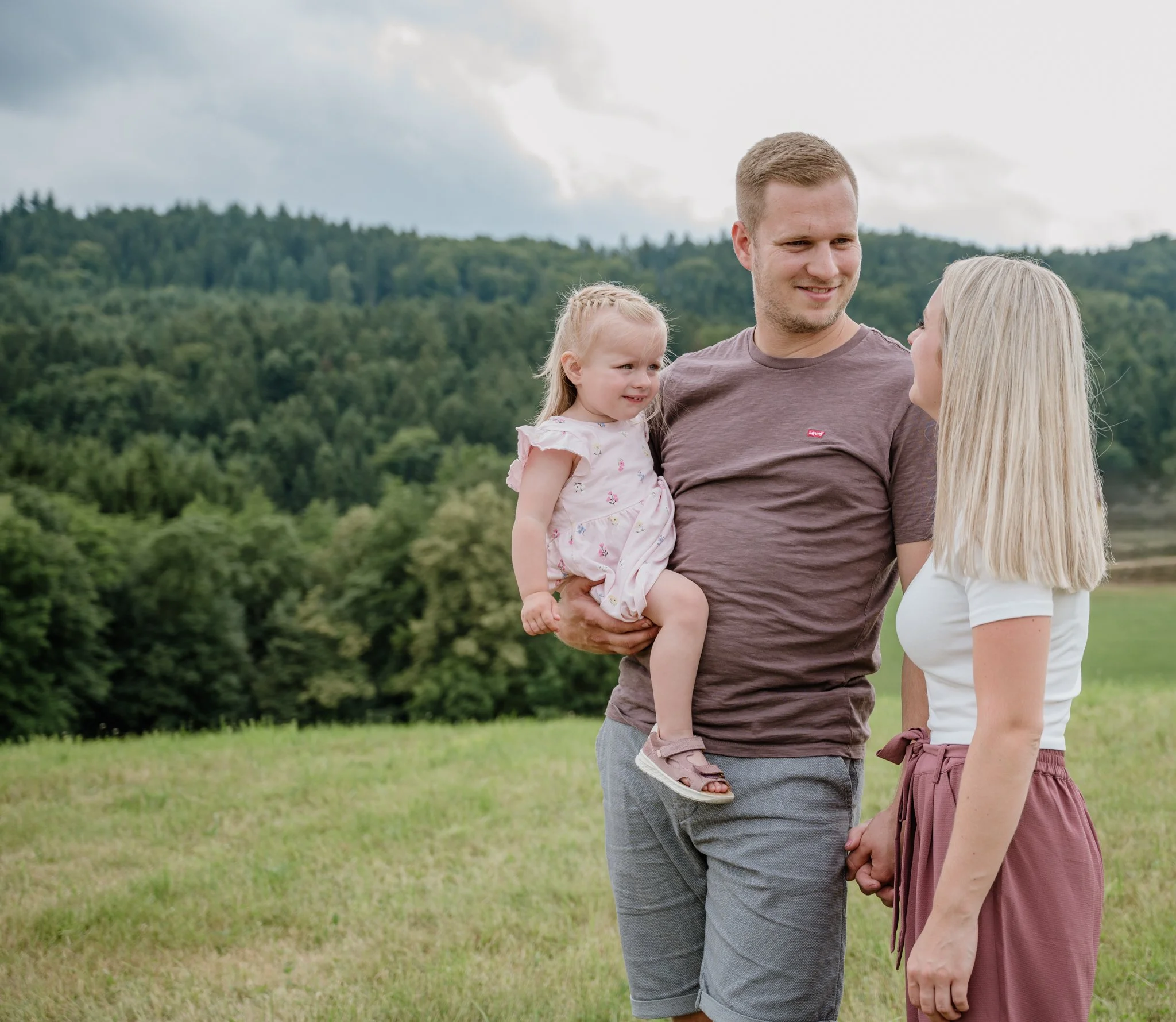 Familie im Freien, ein Mann hält ein kleines Mädchen, ein blonde Frau hält die Hand, umgeben von grüner Landschaft und Hügeln, bewölkter Himmel im Hintergrund.