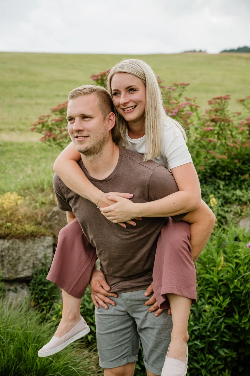 Ein junges Paar, das draußen in der Natur ist, wobei der Mann eine Frau auf seinem Rücken trägt. Beide lächeln und wirken glücklich, umgeben von grünen Pflanzen und einem hügeligen Hintergrund.