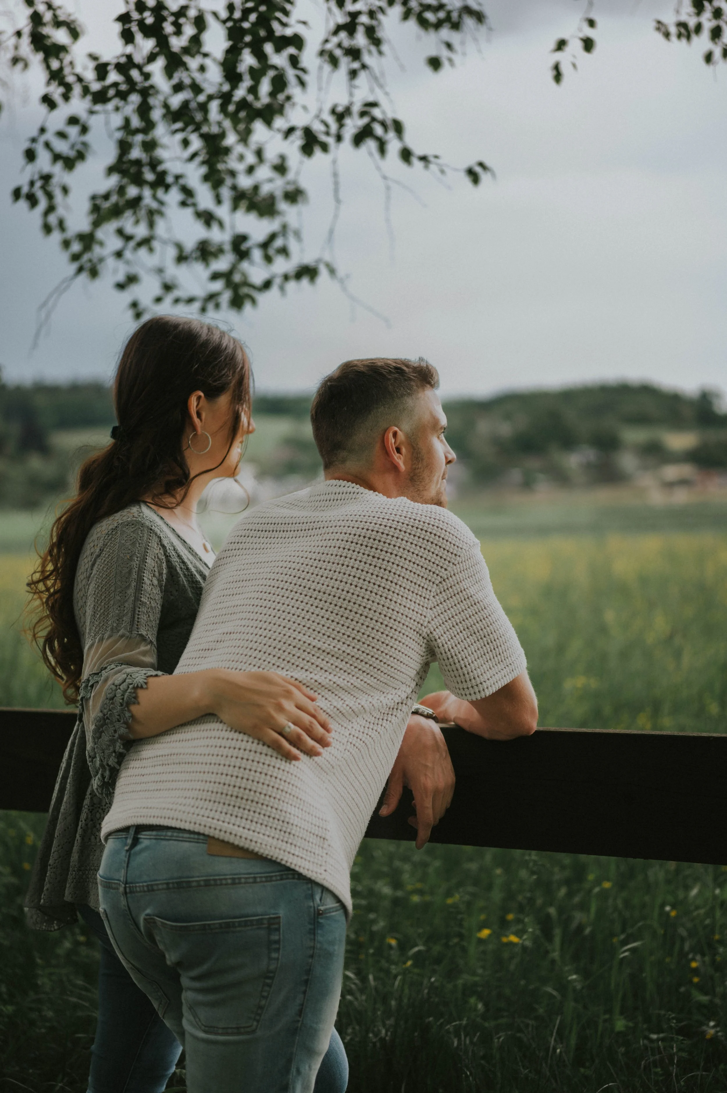 Ein Paar steht an einem Zaun und blickt in die Landschaft, umgeben von grünen Feldern und Bäumen.
