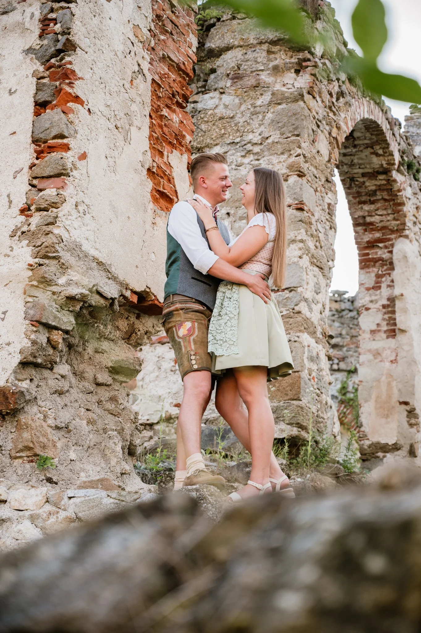 Ein Paar umarmt sich vor den Ruinen einer alten steinernen Burg, lachend und glücklich.