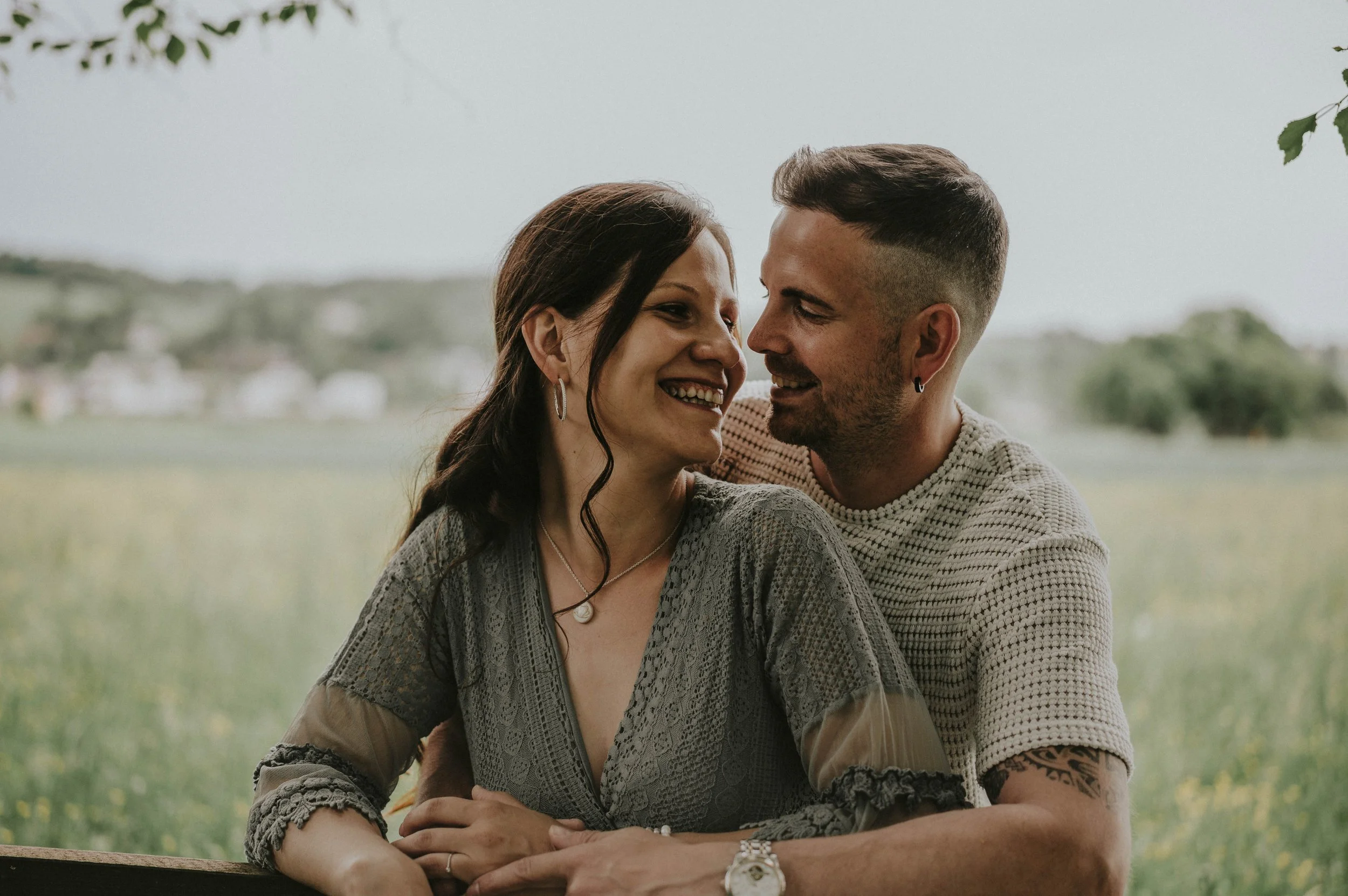 Ein lächelndes Paar sitzt eng beieinander im Freien, umgeben von Natur mit grünen Bäumen und einem Feld im Hintergrund.