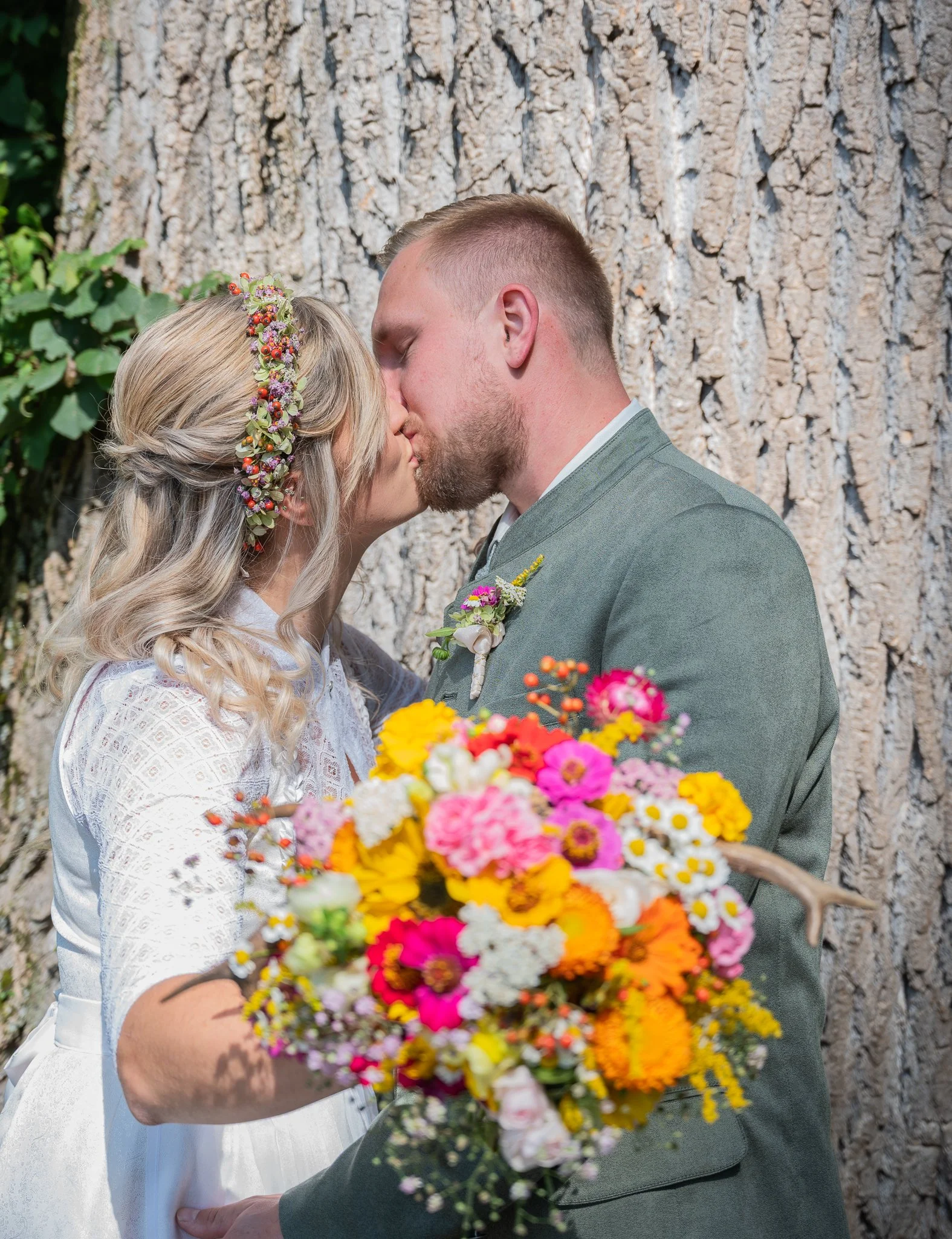 Ein Brautpaar bei einer Hochzeitszeremonie,sektenfreundlich, die Braut hält einen bunten Blumenstrauß, beide küssen sich vor einem Baum.