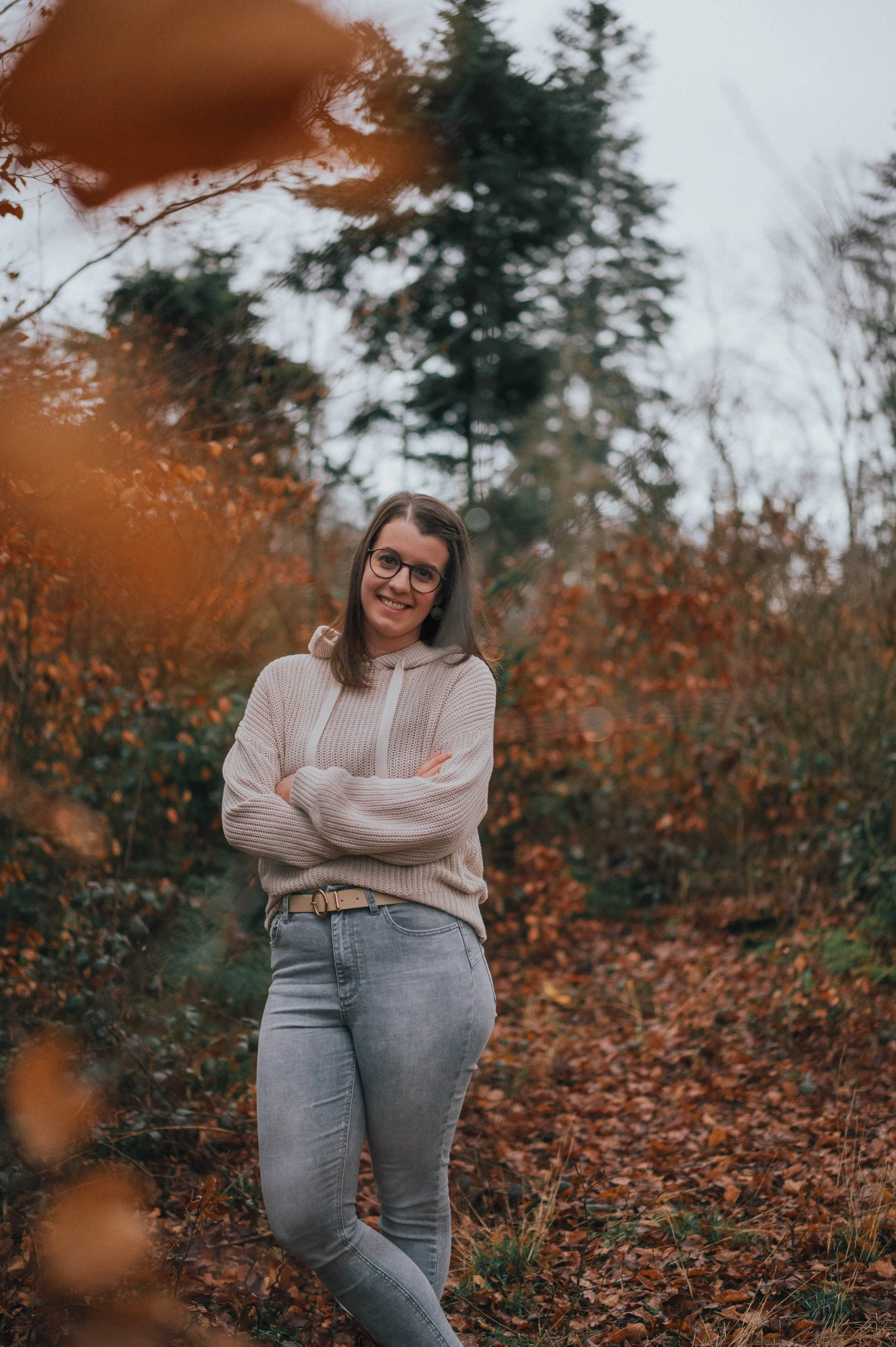 Eine lächelnde Frau mit Brille steht im Herbstlaub in einem Wald, trägt eine beige Strickjacke und graue Jeans.