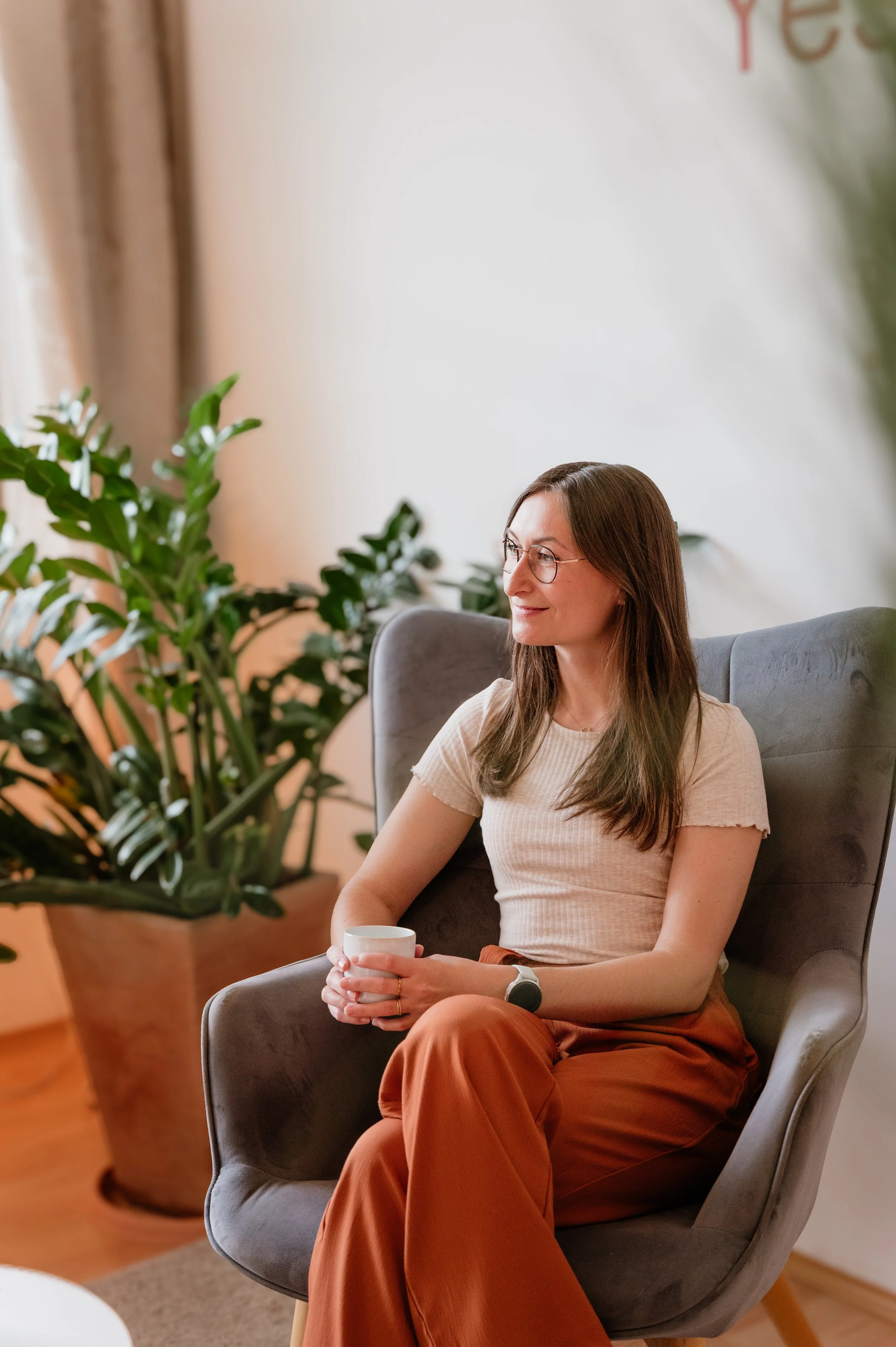 Eine Frau mit Brille sitzt entspannt in einem gemütlichen Raum, hält eine Tasse in den Händen und schaut grübellos zur Seite.