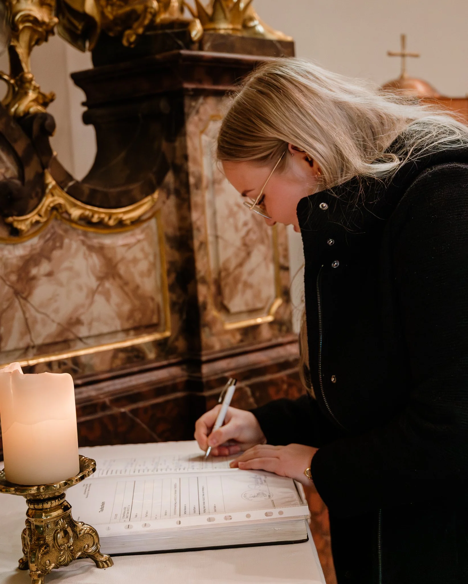 Eine Frau schreibt in ein Buch auf einem Tisch, neben einer Kerze in einer goldenen Kerzenhalter, im Hintergrund ein kunstvoller Holz- und Marmorschrank und eine kleine goldene Metallsäule.