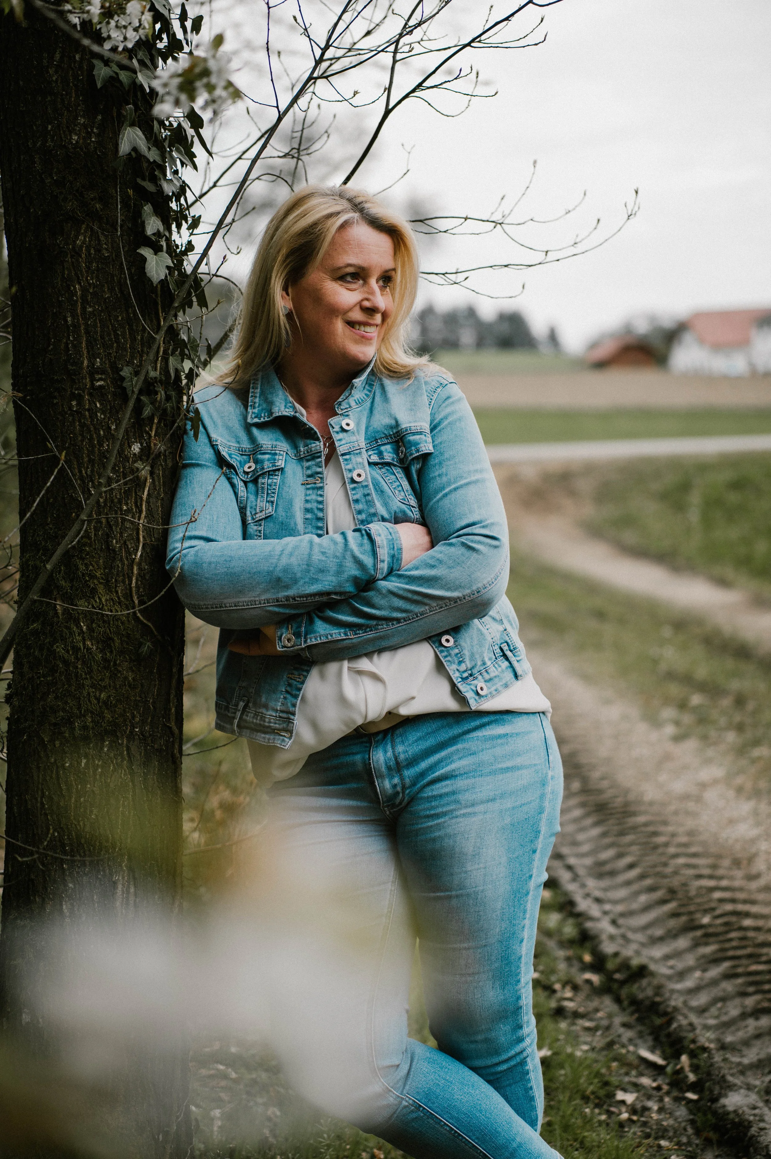 Eine Frau lehnt an einem Baum in einer ländlichen Umgebung mit Feldern und Häusern im Hintergrund, trägt eine Jeansjacke und Jeans, lächelt und blickt nach rechts.