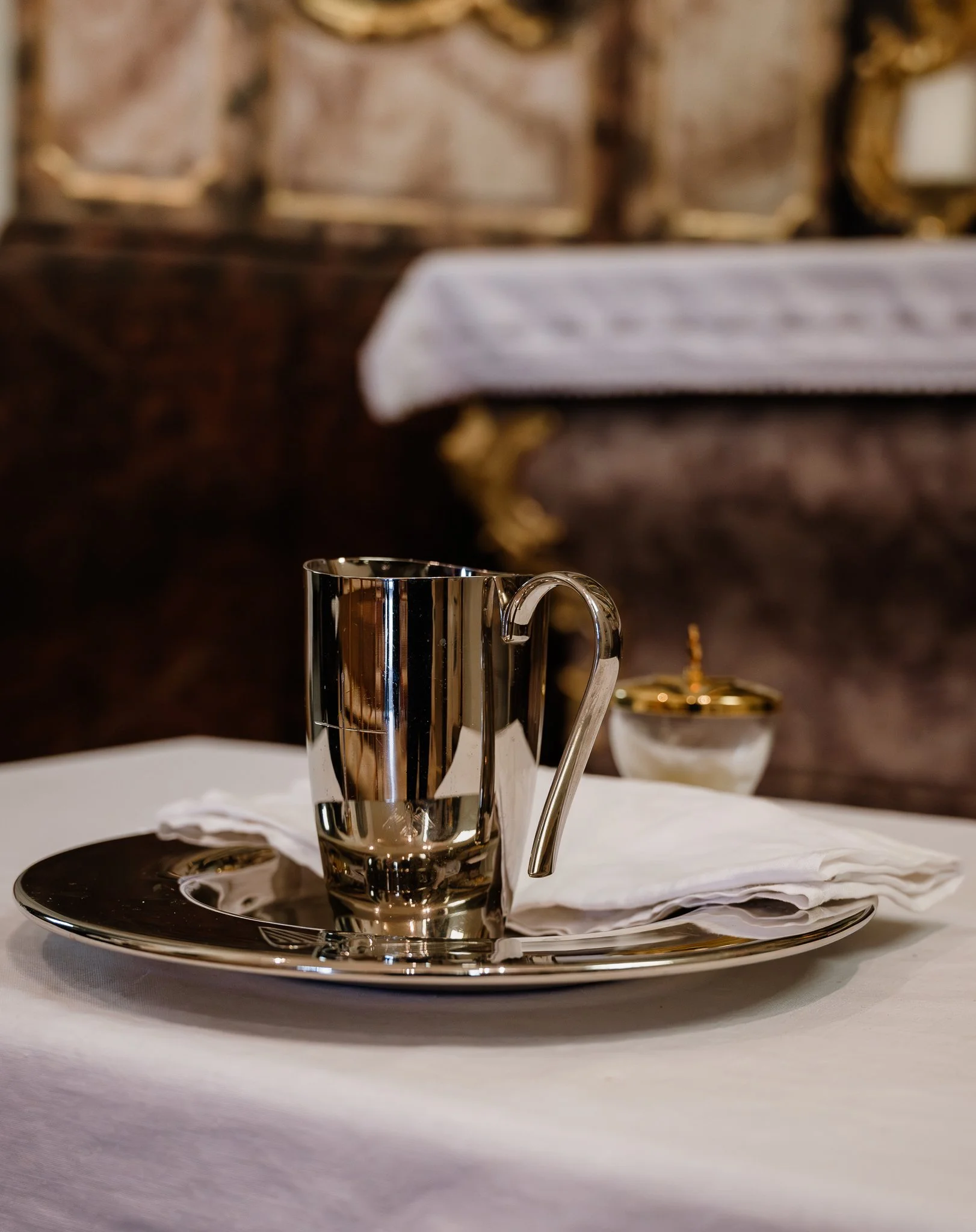 Ein silberner Tassenhalter mit einer Tasse darauf, eine weiße Serviette, auf einem silbernen Teller, auf einem weißen Tuch. Im Hintergrund ein verschnörkelter goldener Rahmen.