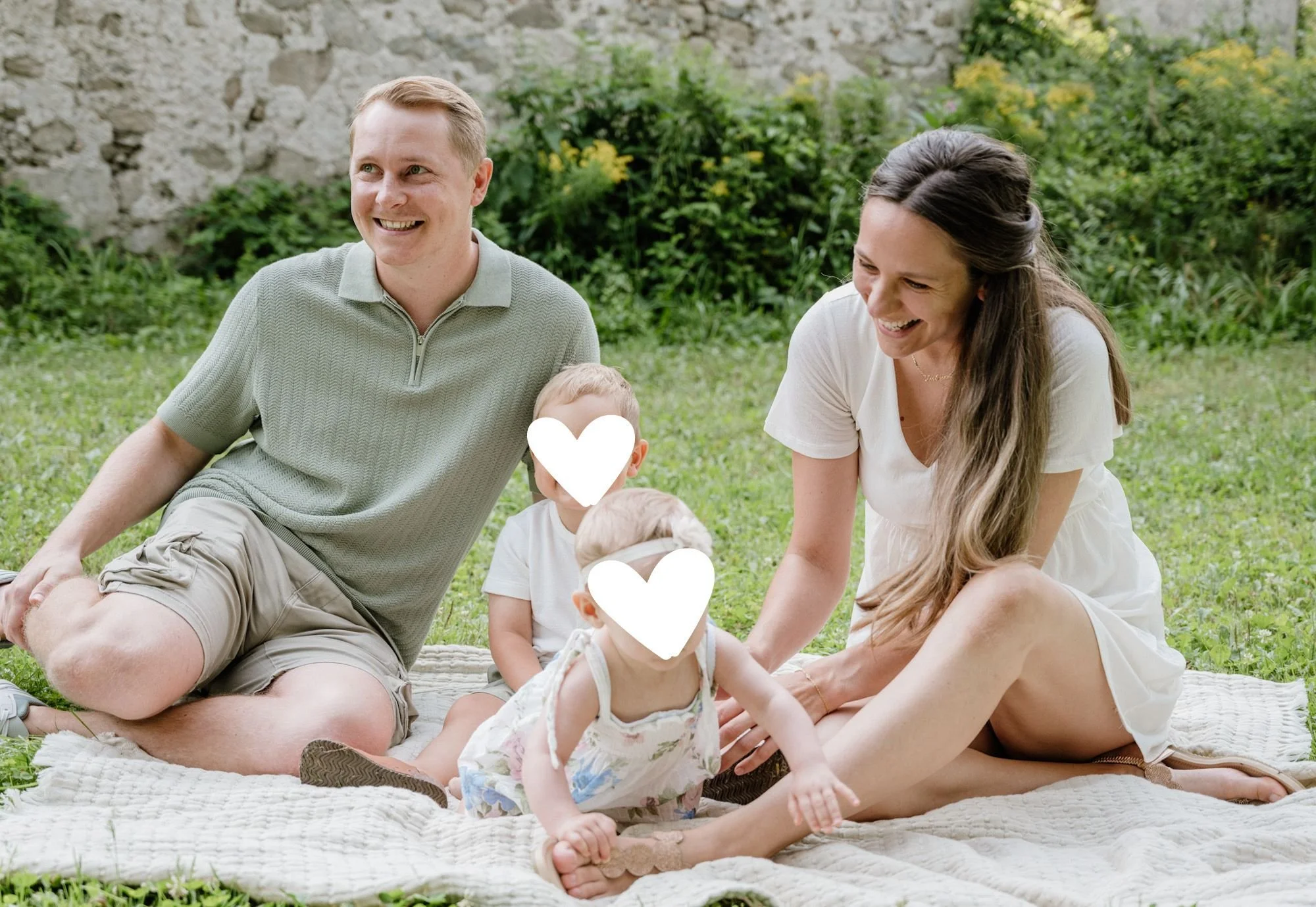 Familie beim Picknick im Garten, sitzend auf einer Decke und lachend, mit zwei kleinen Kindern, Natur im Hintergrund.