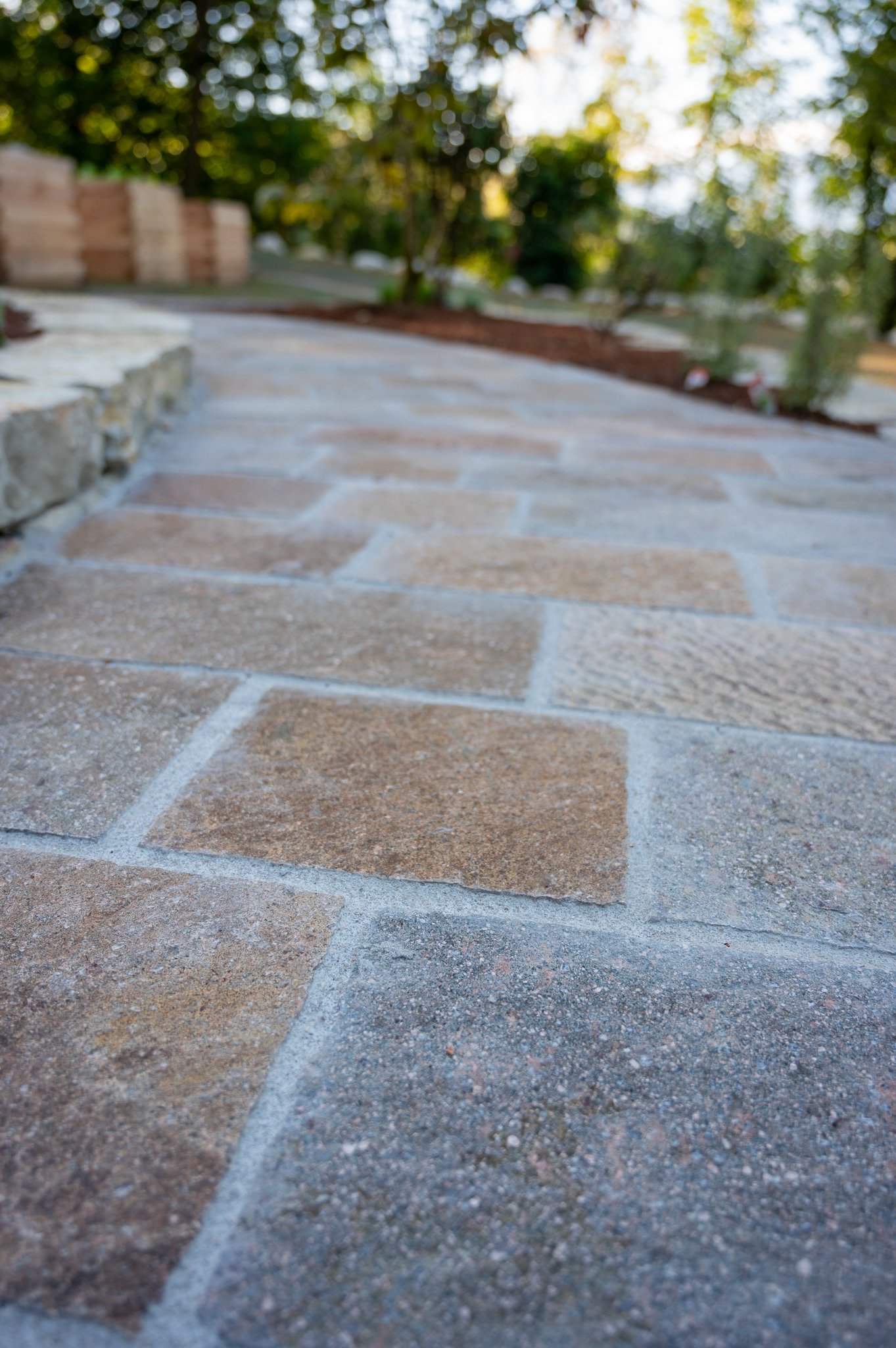 Nahaufnahme eines gepflasterten Weges mit Steinen in natürlichen Farben, umgeben von Bäumen bei Sonnenlicht.