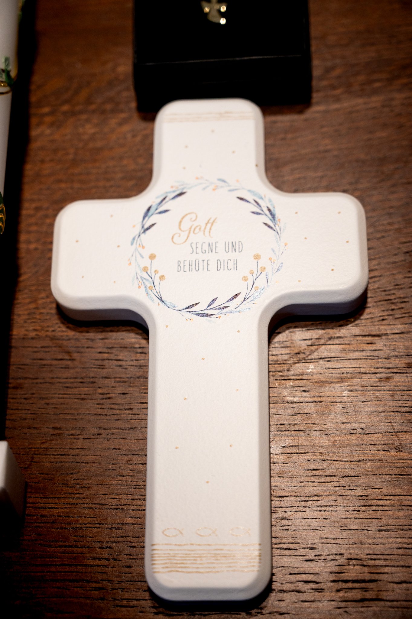 Ein weißes Kreuz mit der Aufschrift 'Gott segne und behüte dich' auf einer Holzoberfläche.