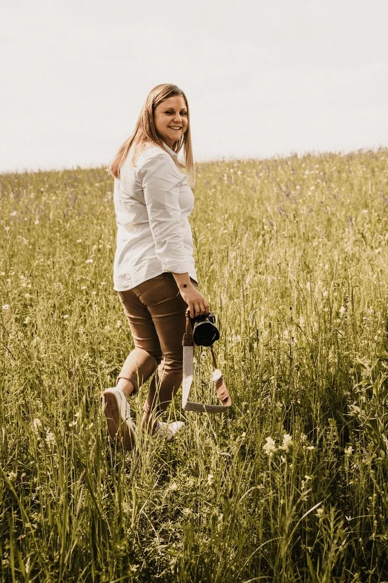 Frau steht in einem grünen Blütenfeld, hält eine Kamera in der Hand und lächelt.