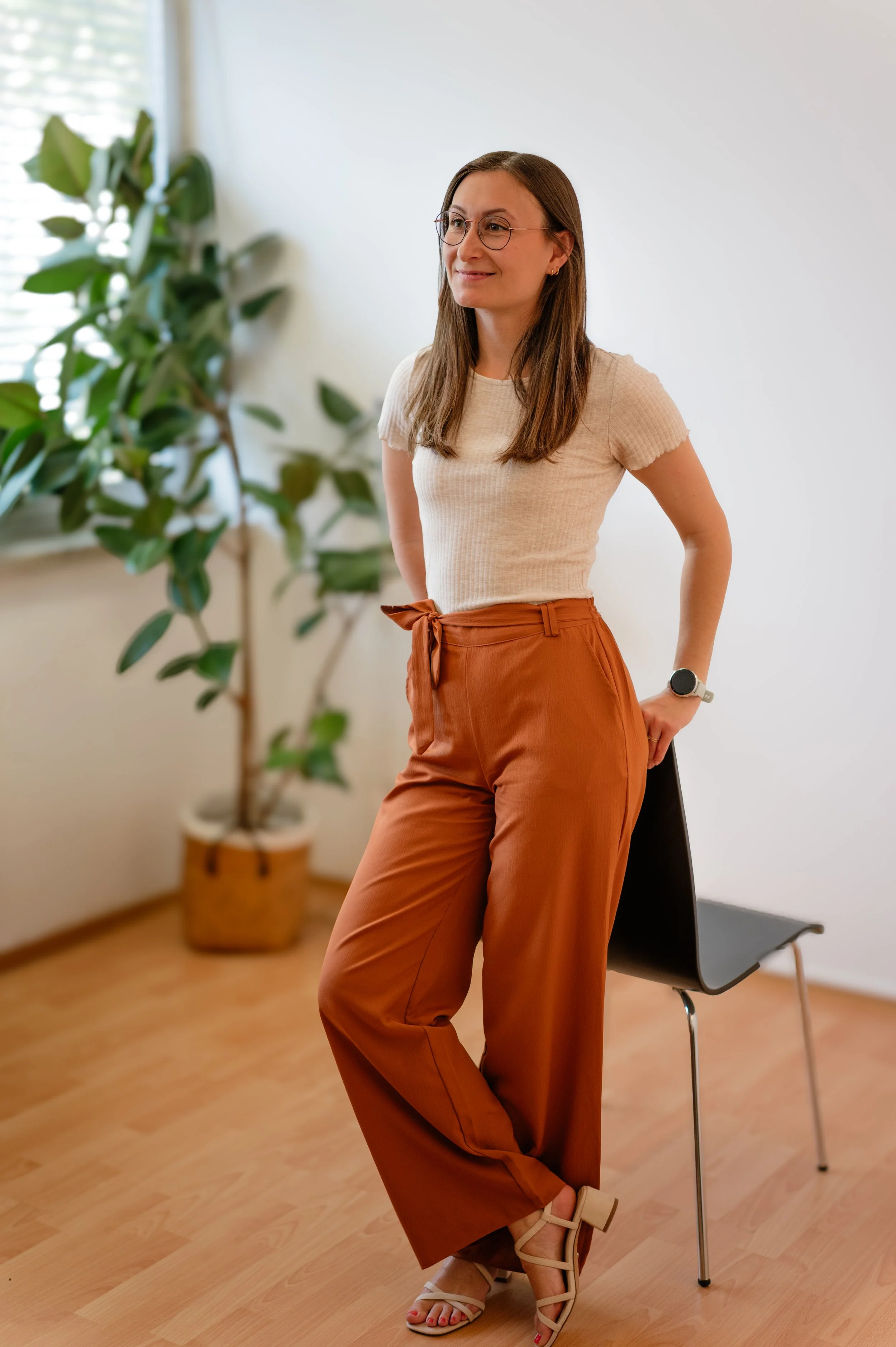 Eine Frau mit Brille, braunen Haaren, in einem beige T-Shirt und braunen, weiten Hosen, steht in einem Raum mit Holzboden und einem weißen Wand, neben einem schwarzen Stuhl und einer großen grünen Pflanze.