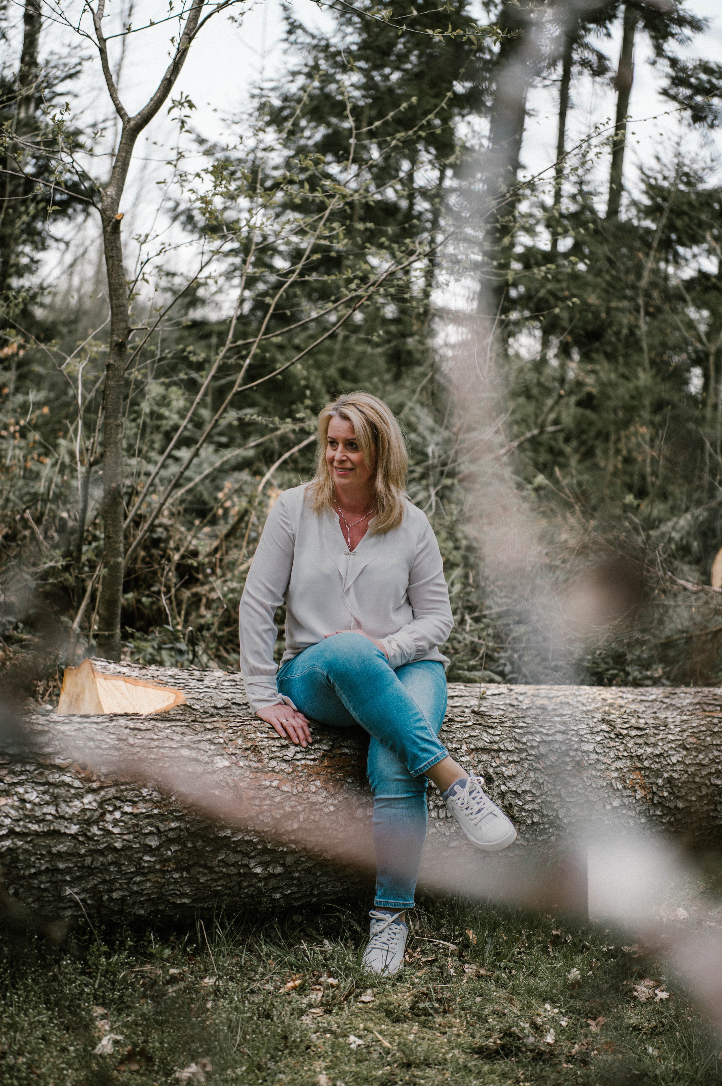 Eine Frau sitzt auf einem Baumstamm in einem Wald, umgeben von Bäumen und Zweigen.