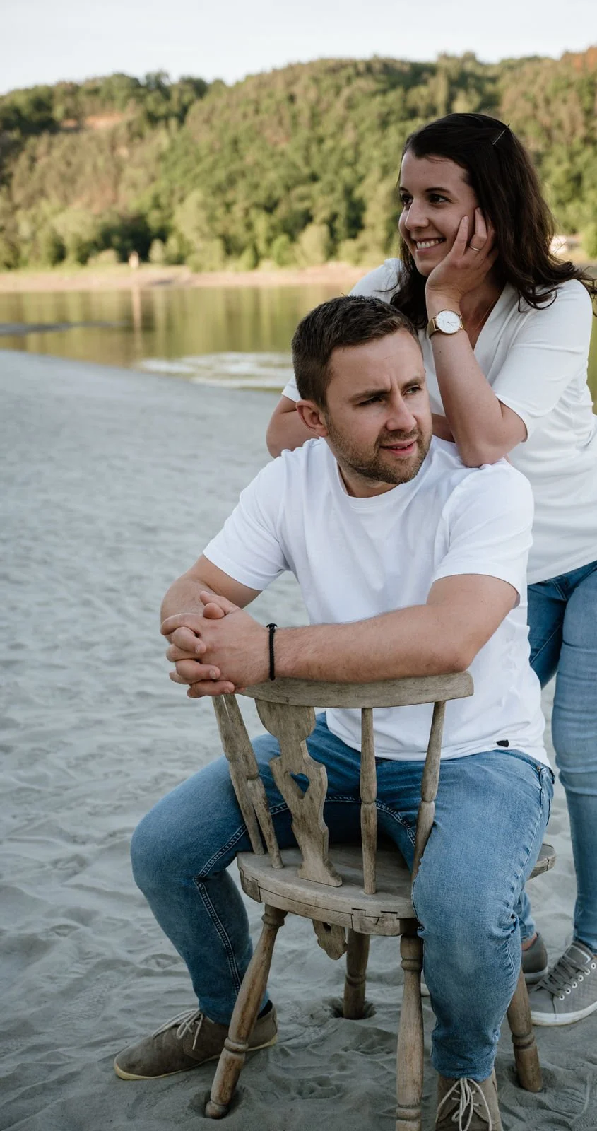 Ein Mann sitzt auf einem Holzstuhl im Sand, während eine Frau hinter ihm sitzt und ihn freundlich ansieht, neben einem See mit bewaldeten Hügeln im Hintergrund.