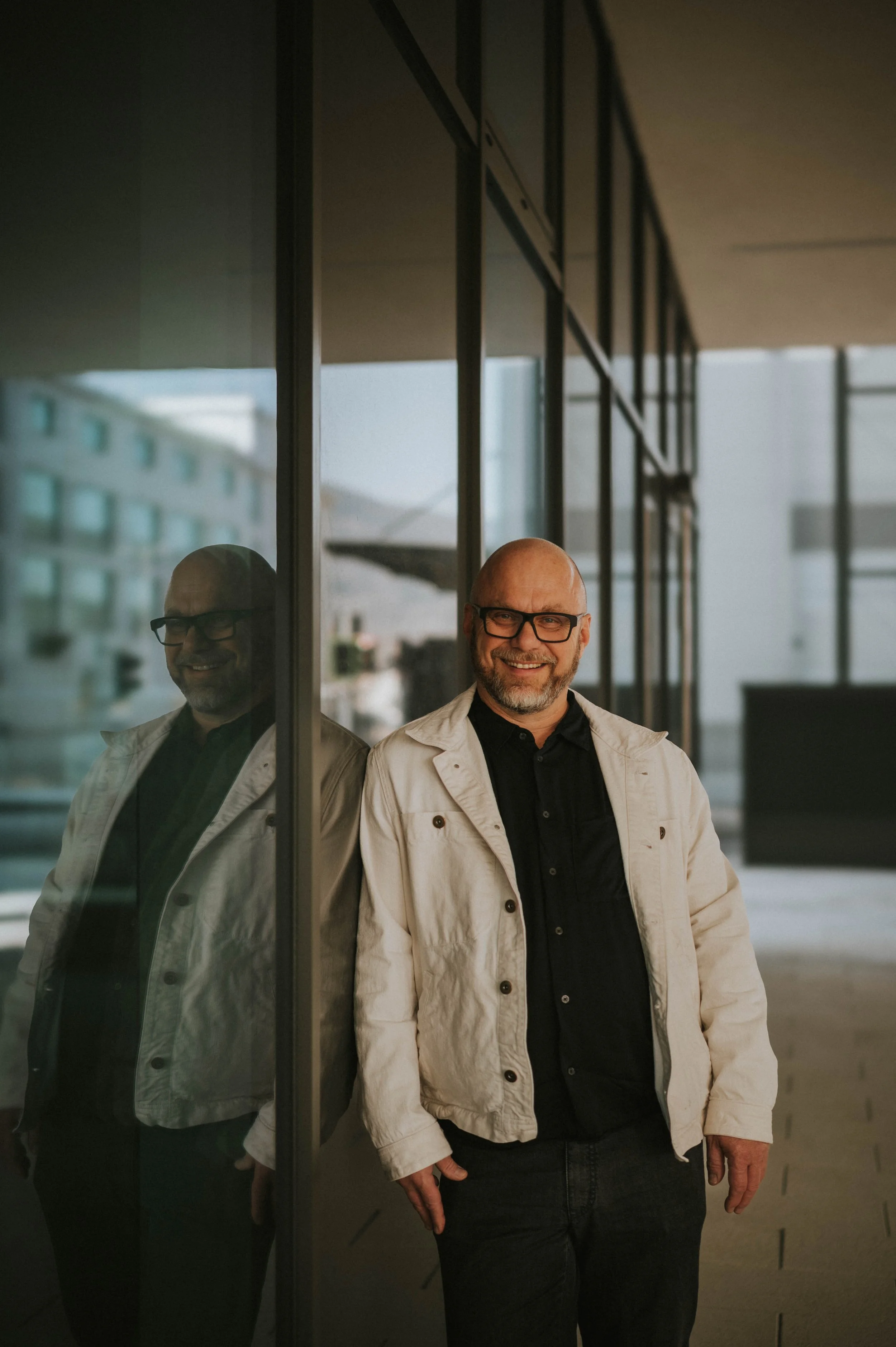 Ein lächelnder Mann mit Brille in einer weißen Jacke lehnt an einer Glaswand, reflektiert im Fenster, im Hintergrund ein modernes Gebäude.