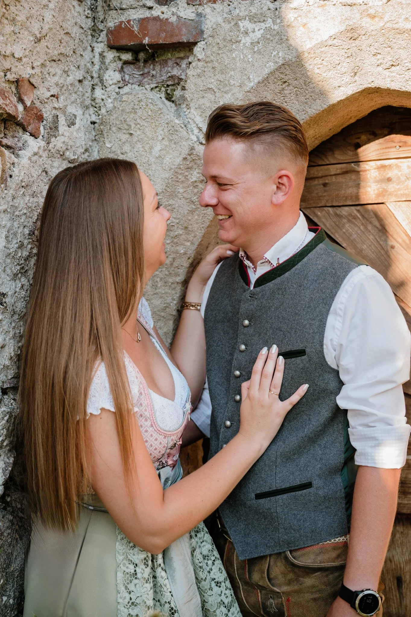 Ein Paar in traditioneller bayerischer Kleidung, die sich in einer alten Steinmauer nähern und glücklich lachen.