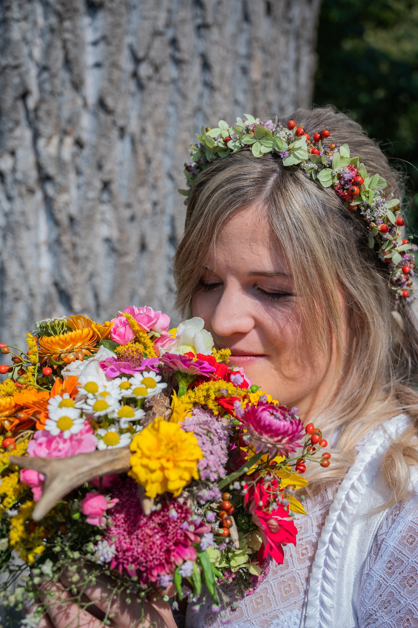 Eine junge Frau trägt einen Blumenkranz auf dem Kopf und hält einen bunten Blumenstrauß nah ans Gesicht, vor einem Baum.
