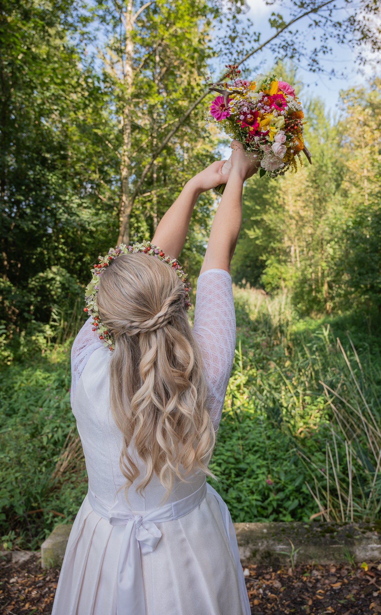 Ein Mädchen in weißer Kleidung mit Blumenkranz auf dem Kopf hält einen bunten Blumenstrauß in die Luft, während sie im Freien steht, umgeben von Bäumen und grüner Natur.