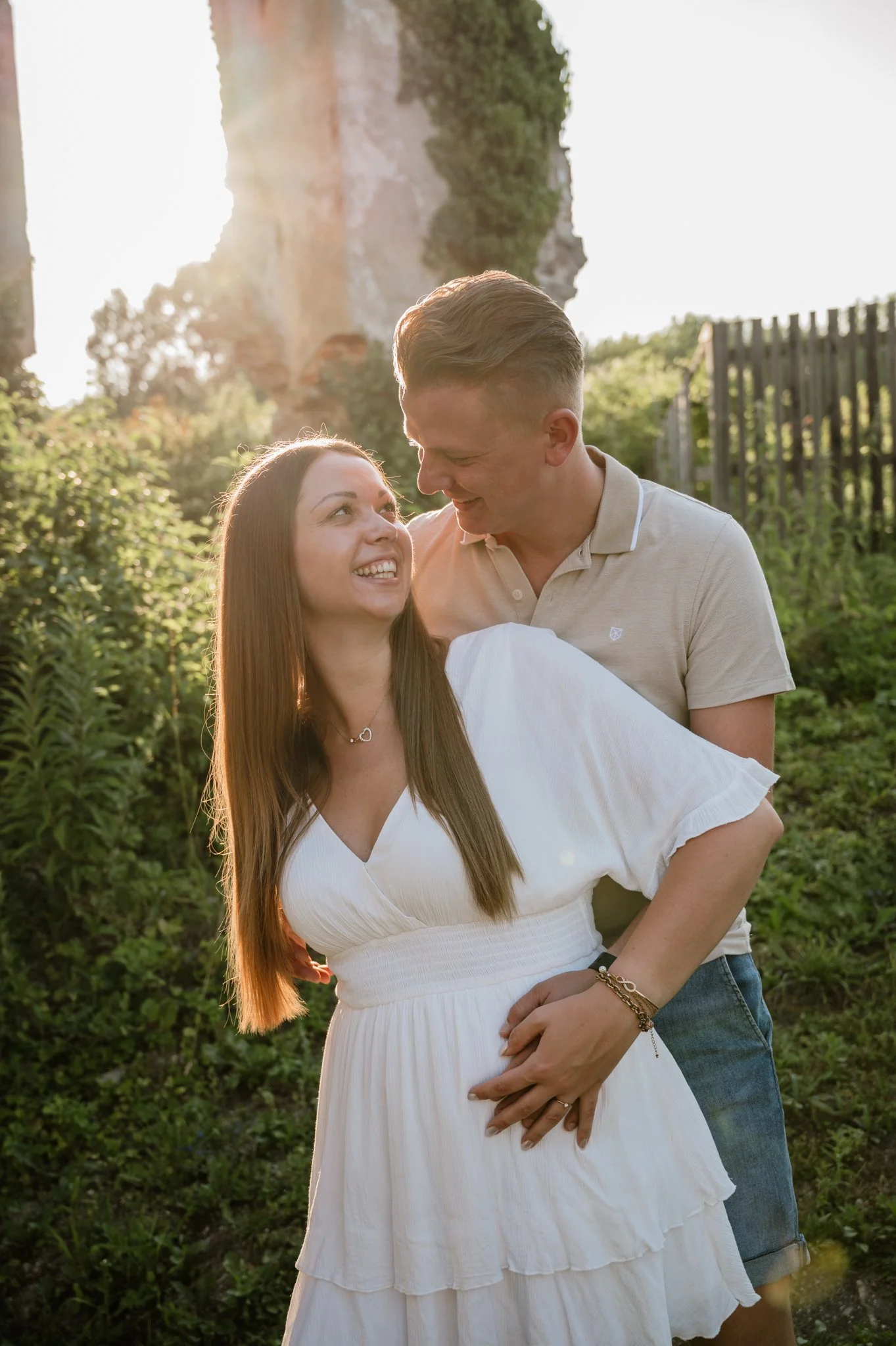 Ein glückliches Paar steht bei Sonnenuntergang im Freien, lächelnd und sich liebevoll anblickend, umgeben von grüner Natur und einem Felsen im Hintergrund.