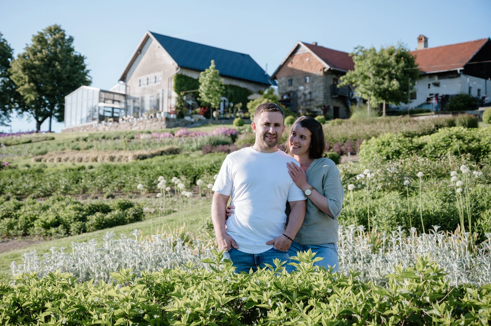 Ein Paar steht in einem Garten mit Blumen und Pflanzen, im Hintergrund sind Häuser auf einem Hügel zu sehen, bei sonnigem Wetter.