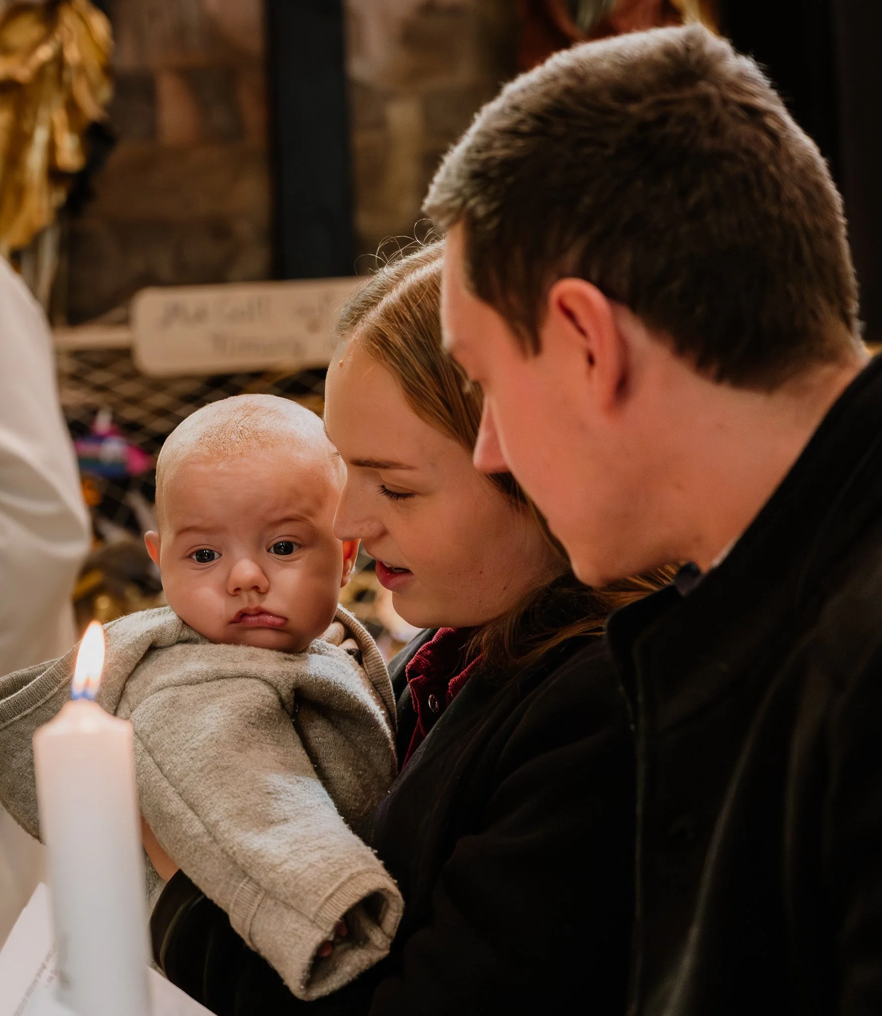 Ein Baby wird von einer jungen Frau und einem jungen Mann in einer Kirche gehalten, während eine Kerze brennt.