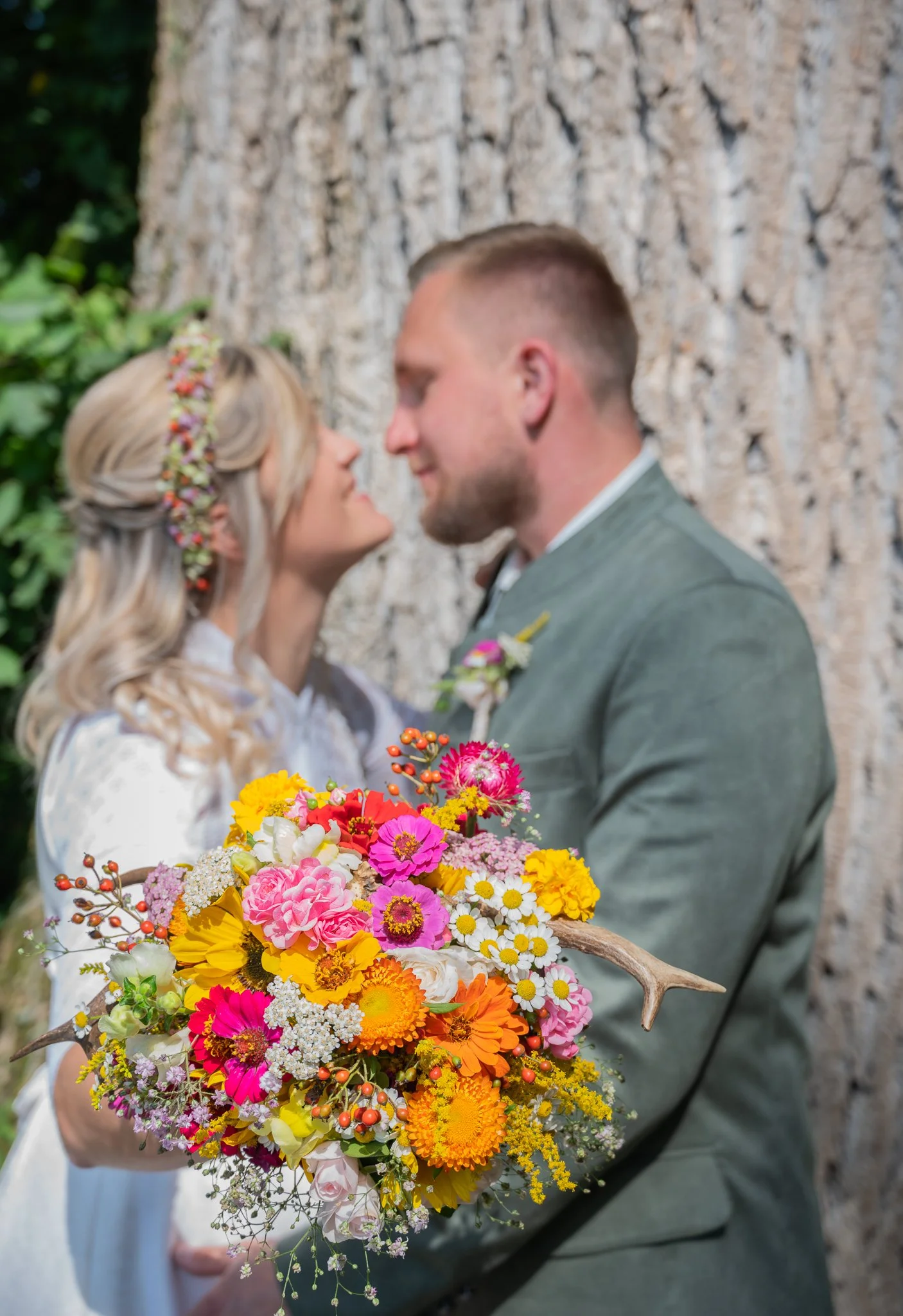 Ein Brautpaar steht eng beieinander vor einem Baum, die Braut hält einen bunten Blumenstrauß.