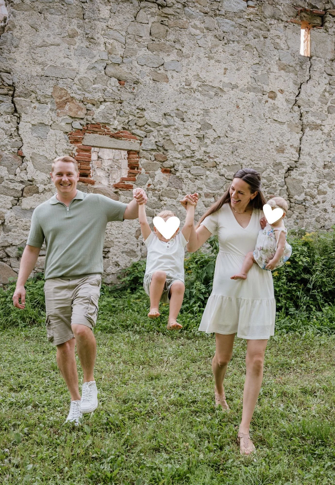 Familie mit zwei Erwachsenen und zwei Kindern, die im Garten vor einer alten Steinmauer Spaß haben. Die Eltern halten die Kinder an den Händen, während eines der Kinder schwingt.