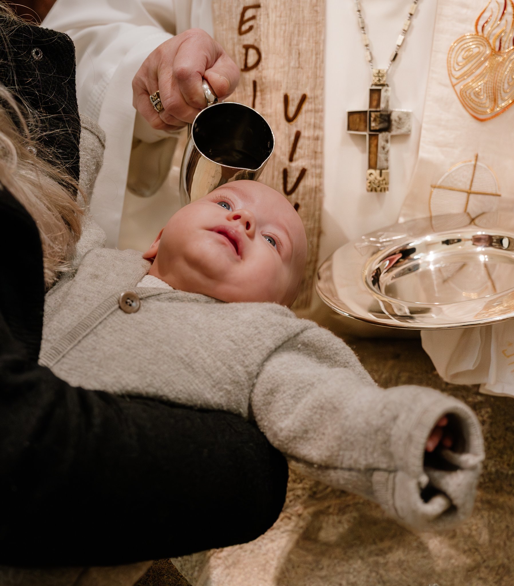 Ein Baby wird während einer religiösen Zeremonie mit Wasser über das Gesicht gesprenkelt, während eine Person im Hintergrund eine Metallkanne hält. Kleidung und religiöse Gegenstände sind sichtbar.