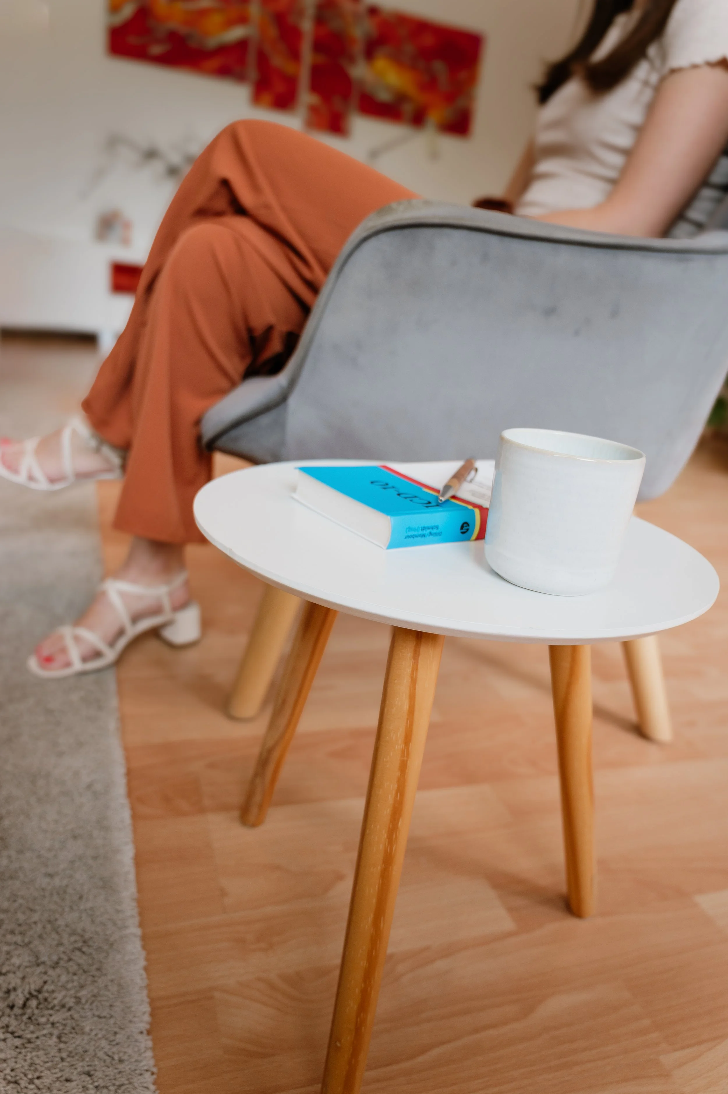 Ein weißer Becher, ein Buch mit blauer Rückseite und ein Kugelschreiber auf einem kleinen weißen Tisch mit Holzfüßen, im Hintergrund eine sitzende Person in weißen Schuhen und roten Hosen, ein grauer Sessel und bunte Wanddekoration.