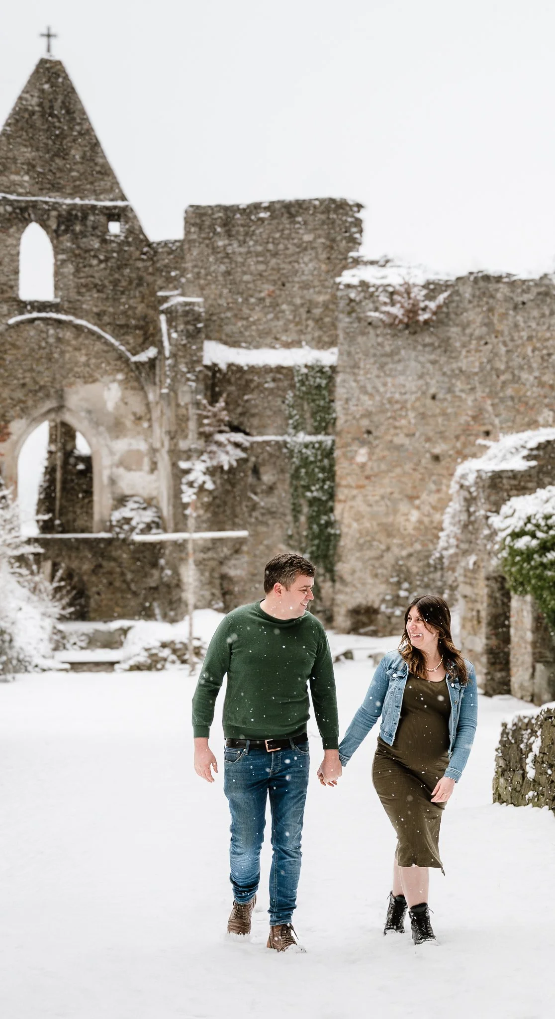 Ein Paar läuft Hand in Hand durch eine verschneite Ruinenlandschaft, im Hintergrund alte, zerfallene Mauern und ein Kirchturm.