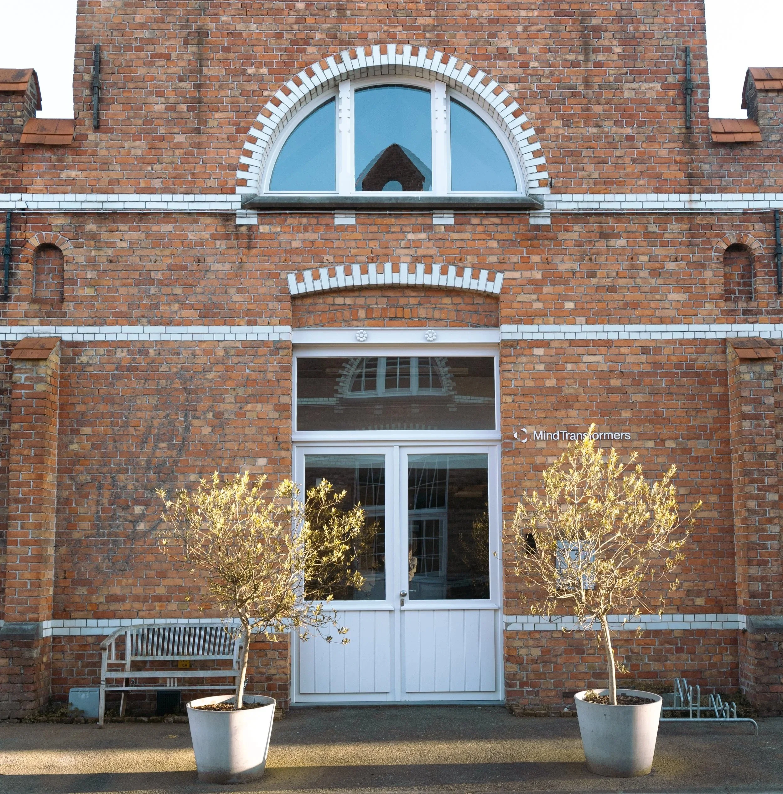 Een bakstenen gebouw met grote ramen, twee kleine bomen in witte potten voor de ingang en een bank naast de linkerboom. Op de muur rechts is de tekst 'Mind Transformers' te zien.