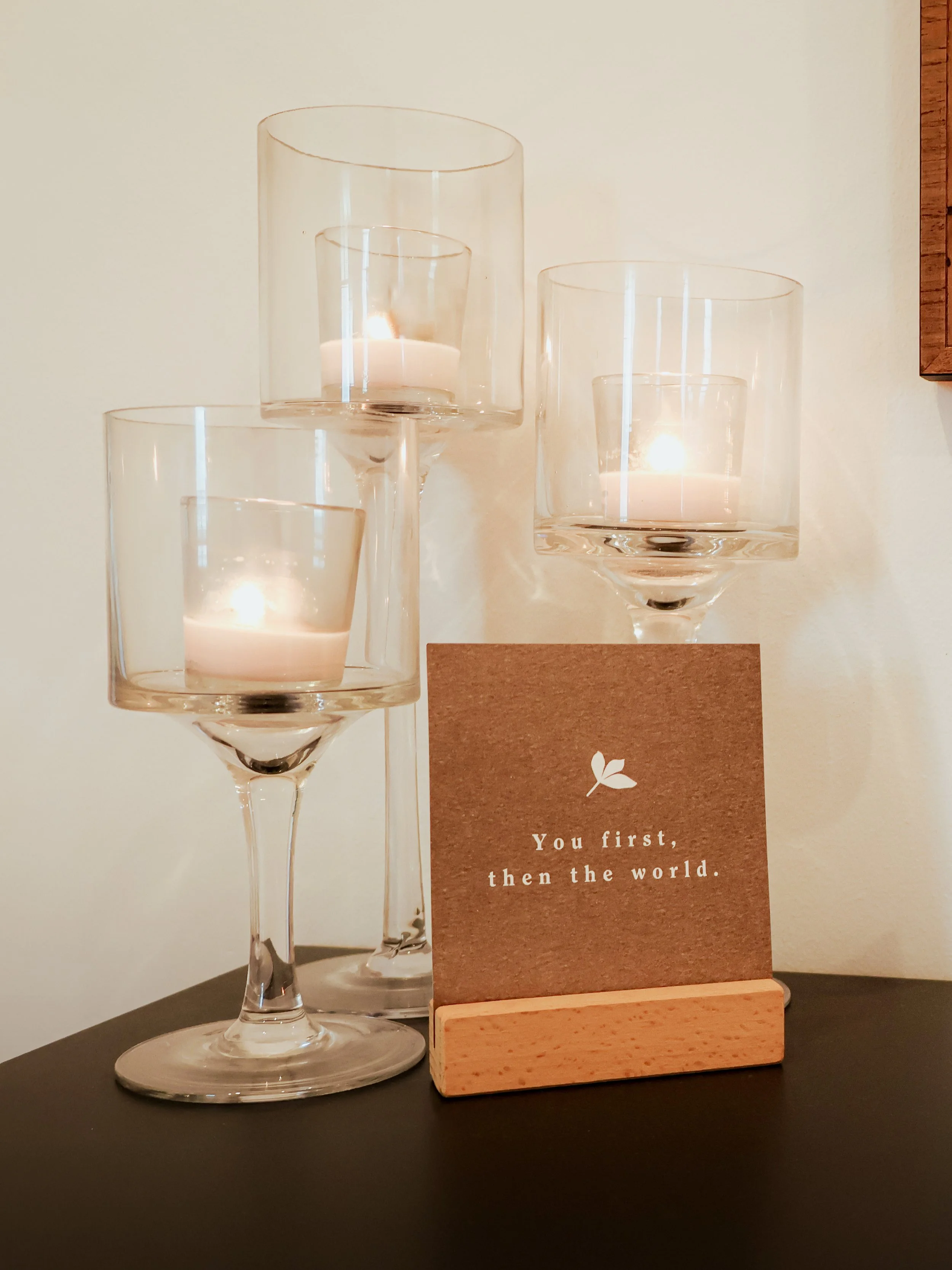 Three glass candle holders with lit candles, a brown card with the quote 'You first, then the world,' and a wooden stand, all placed on a black surface against a light-colored wall.