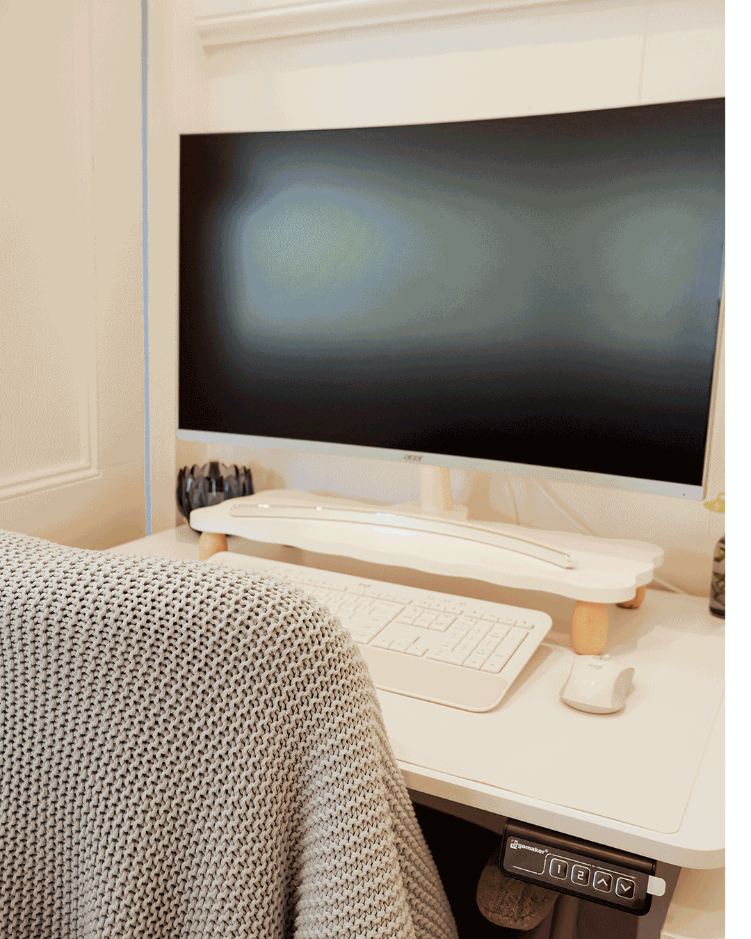 A white computer desk with a large monitor, keyboard, and mouse. There is a knitted blanket on the back of a chair in the foreground, and a small black object and a potted plant to the right of the monitor.