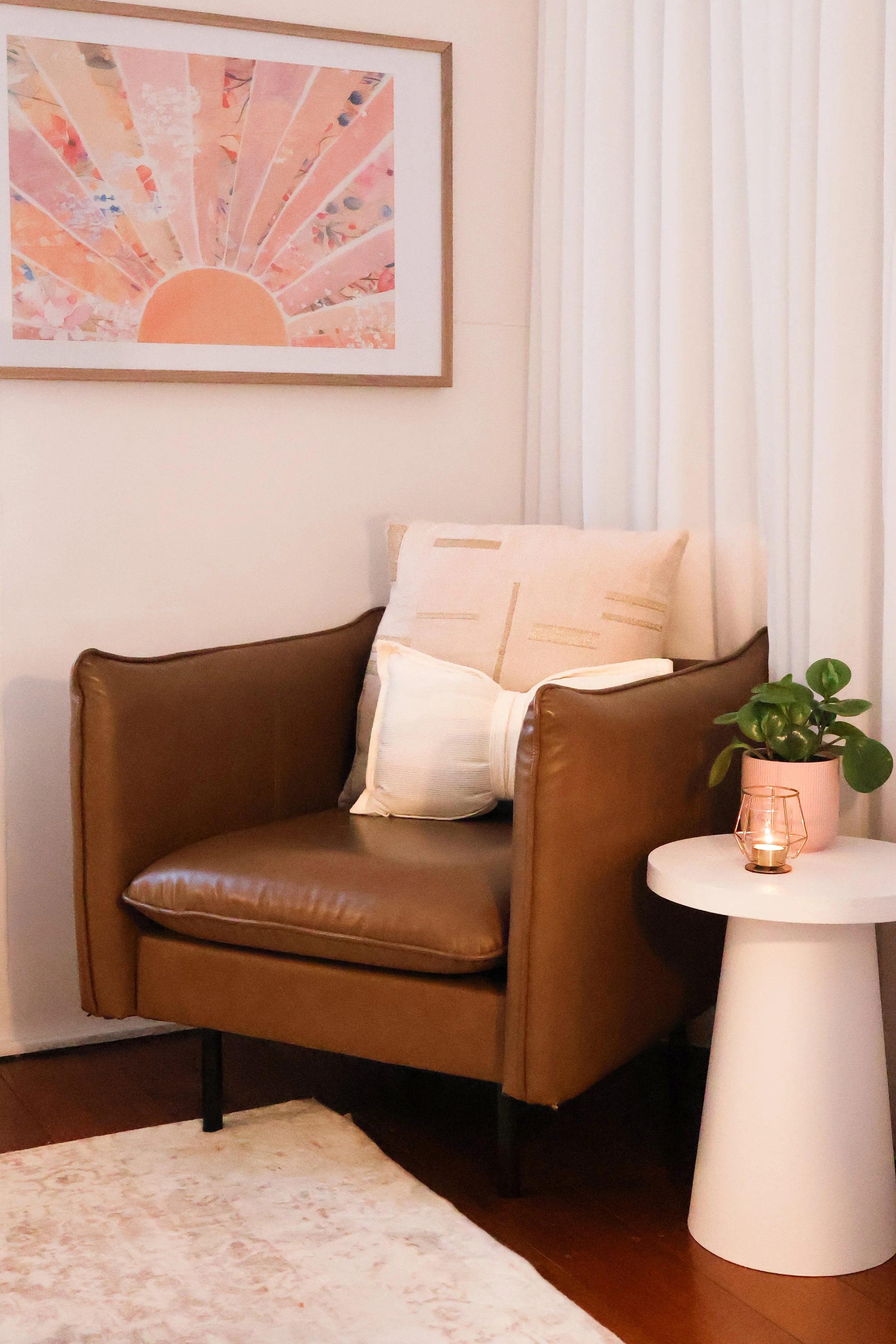 A cozy living room corner with a brown leather armchair, decorated with cream-colored pillows, standing next to a white round side table with a potted plant and a lit candle. Behind the chair, white curtains cover a window, and a framed pink and orange abstract artwork hangs on the wall.