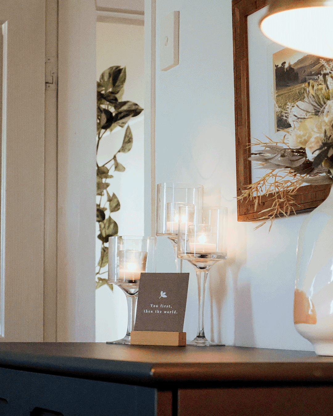 Decorative tabletop with candles in tall glass holders, a small card with the quote 'You first, then the world,' a large framed landscape painting, and floral arrangements in a white vase on a dark wooden surface.