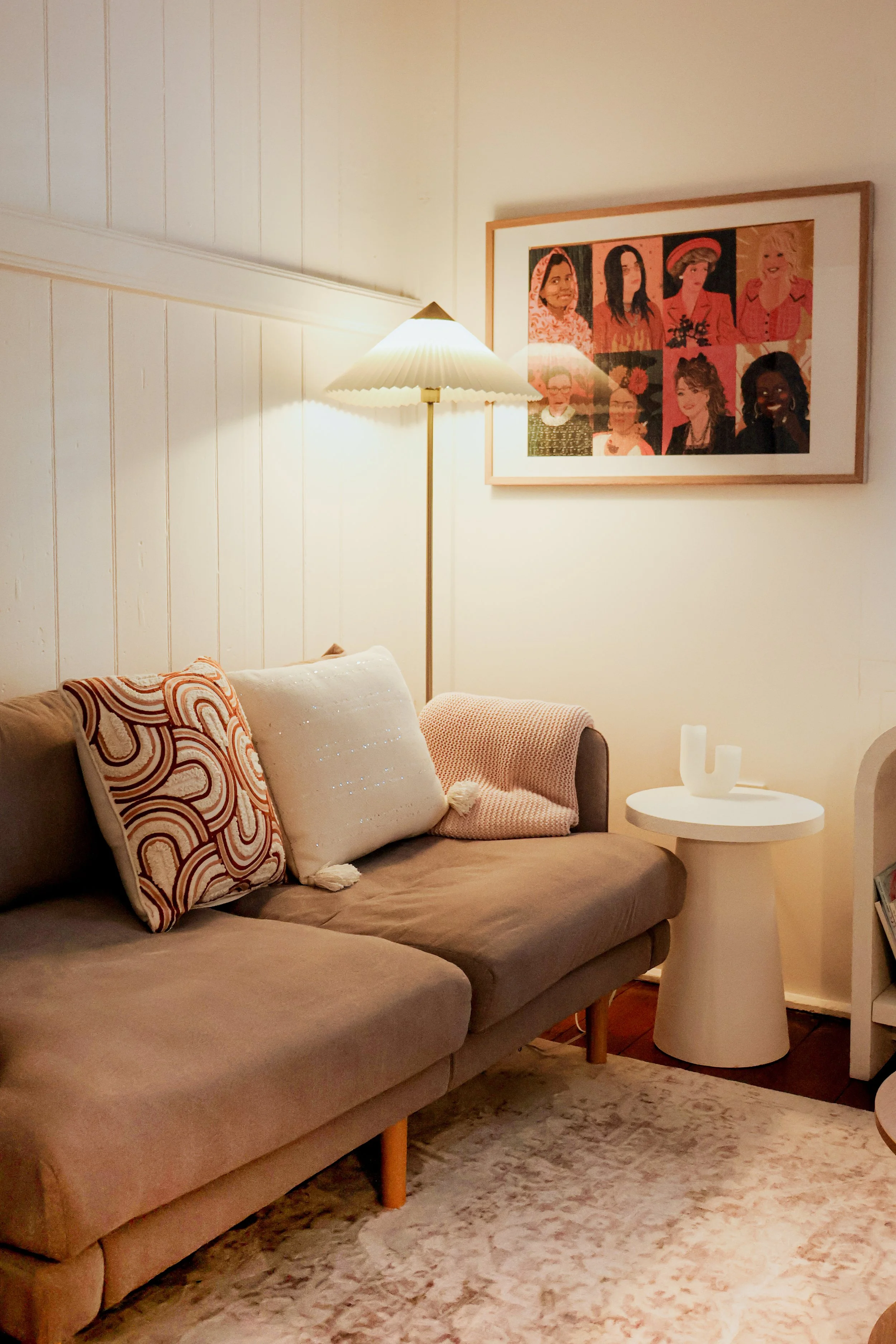A cozy living room with a beige sofa decorated with patterned and plain cushions, a white side table with two candle holders, a framed colorful portrait on the wall, a floor lamp, and a patterned area rug.