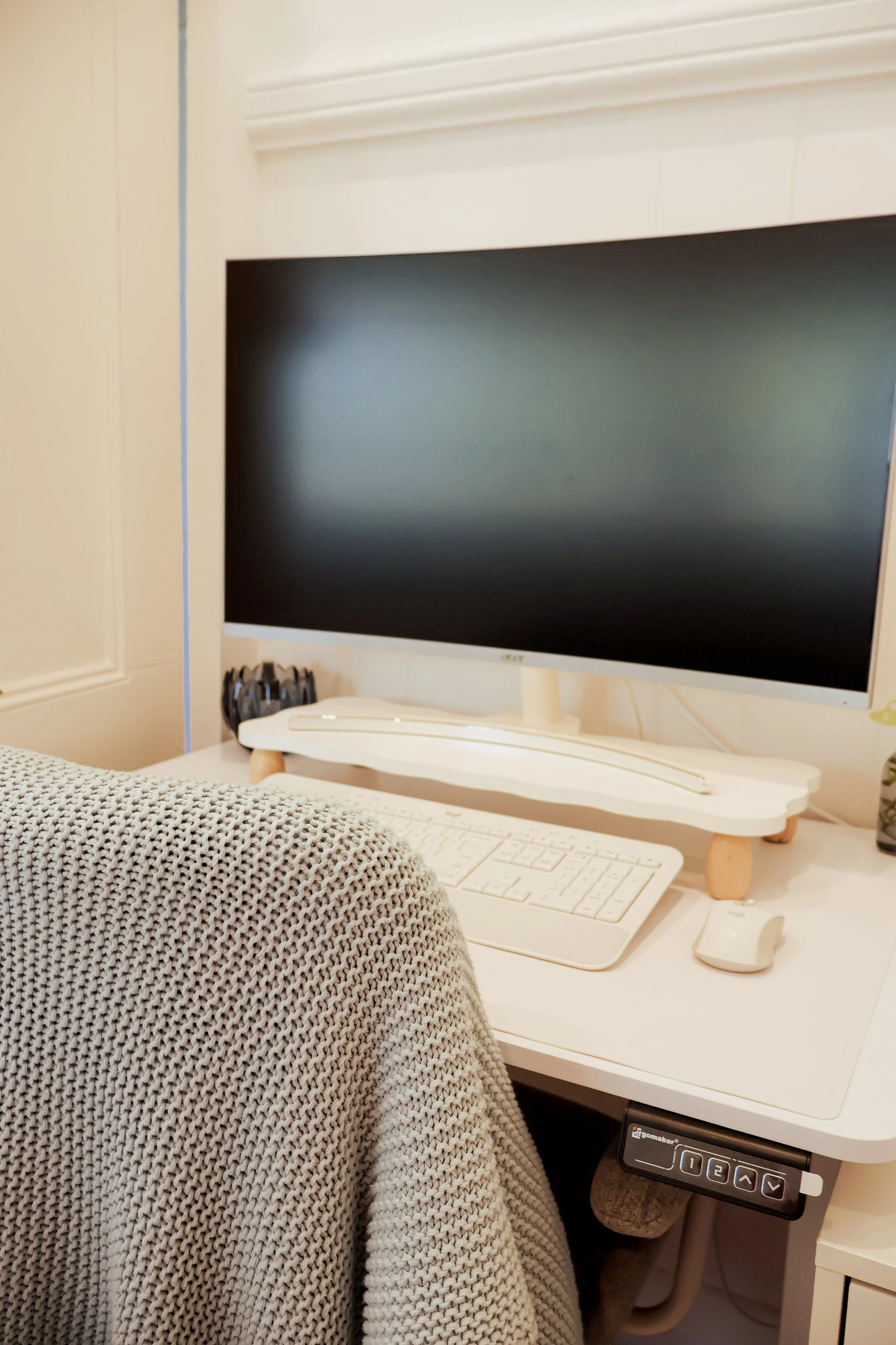 A computer workspace with a large monitor, keyboard, mouse, and a cozy gray sweater draped over a chair in a bright, minimalist room.