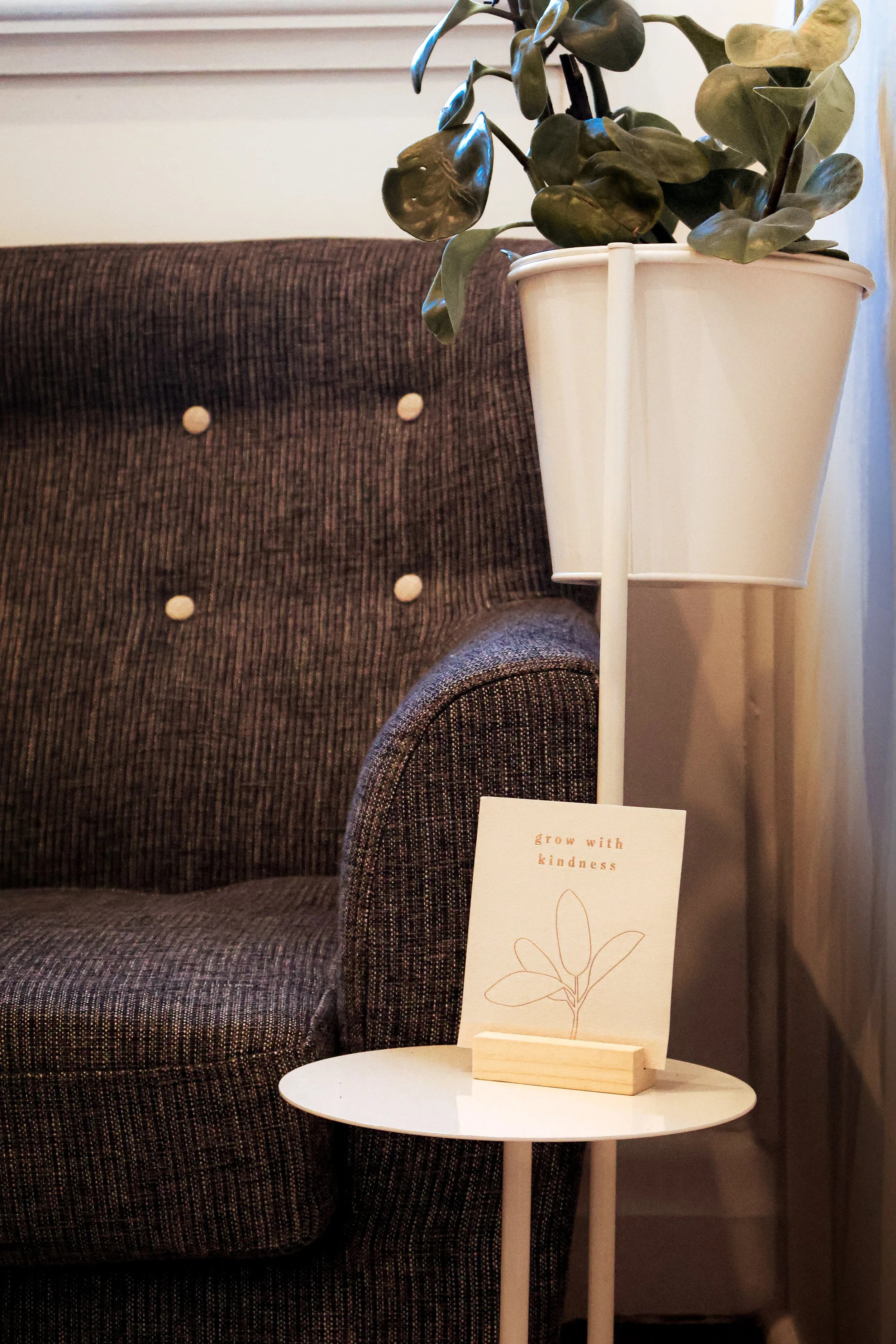 A cozy living space with a dark upholstered chair, a white side table, and a tall white planter with green plant leaves. A small card on the table reads 'grow with kindness' with a simple line drawing of a plant.