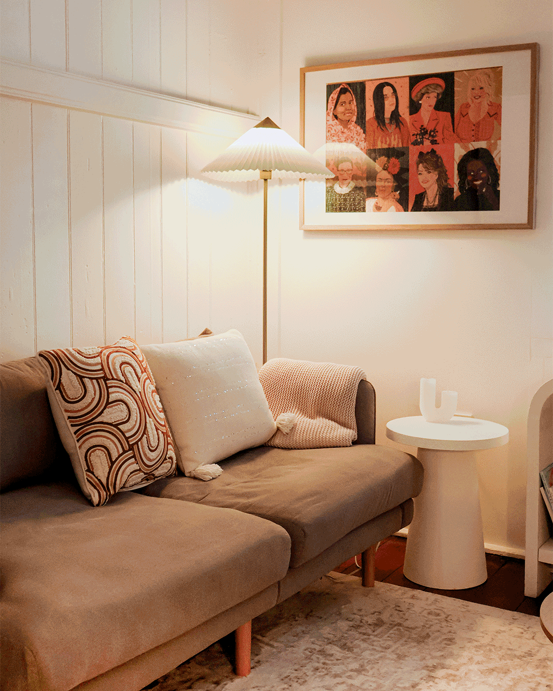 Cozy living room with a beige sofa adorned with patterned and white throw pillows, a floor lamp emitting warm light, and a framed collage of women portraits on the wall.