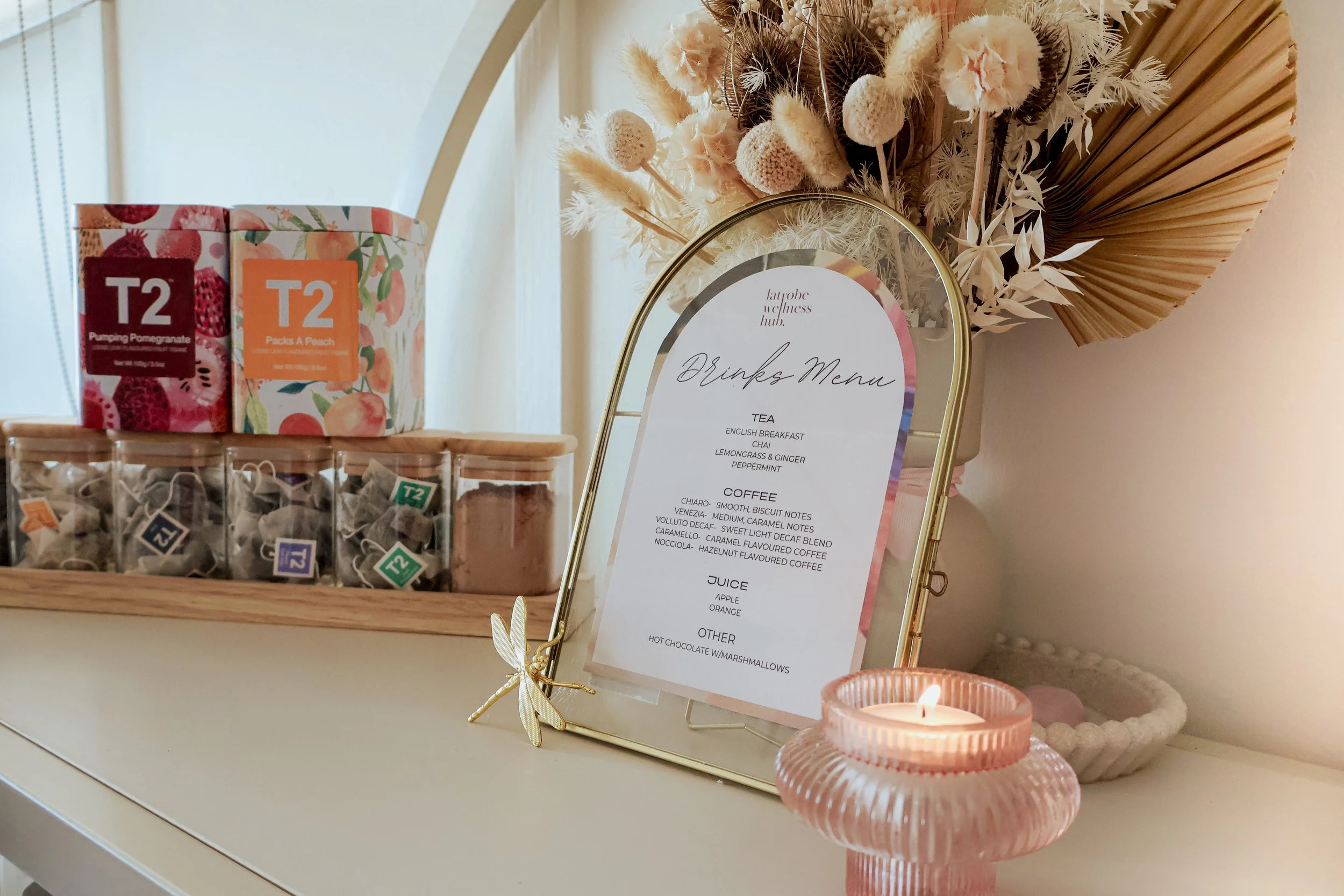 A floral decorative arrangement with dried flowers and leaves. A white framed drink menu with gold border and cursive text. A pink candle holder with a lit candle. Various tea and coffee boxes and loose tea bags on a wooden tray.