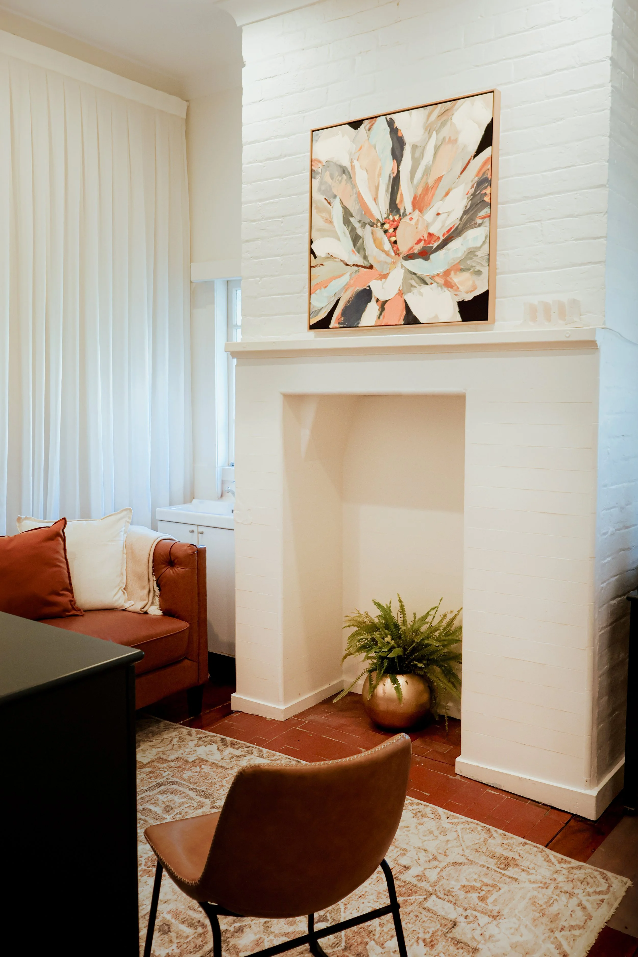 Living room with white brick fireplace, abstract flower painting above, brown leather armchair, sofa with pillows, and green plant in a gold pot.