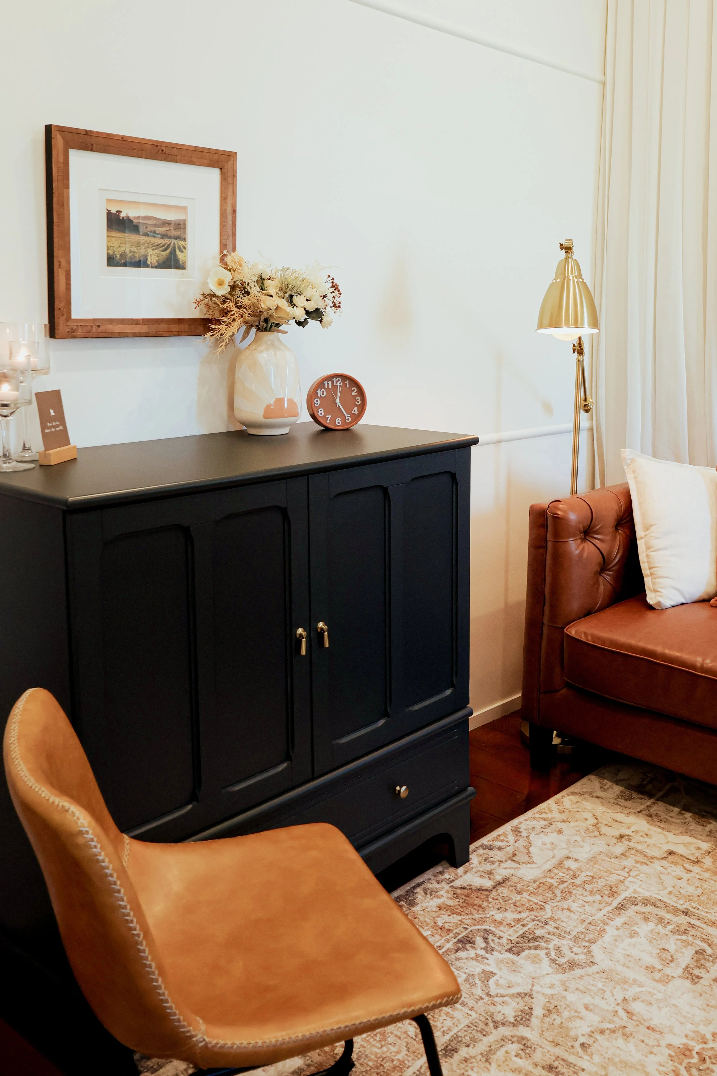 Living room corner with a black cabinet, a white vase with flowers, a round orange clock, a gold floor lamp, a leather armchair, and a brown leather sofa with a white pillow, next to cream curtains and a beige patterned rug.