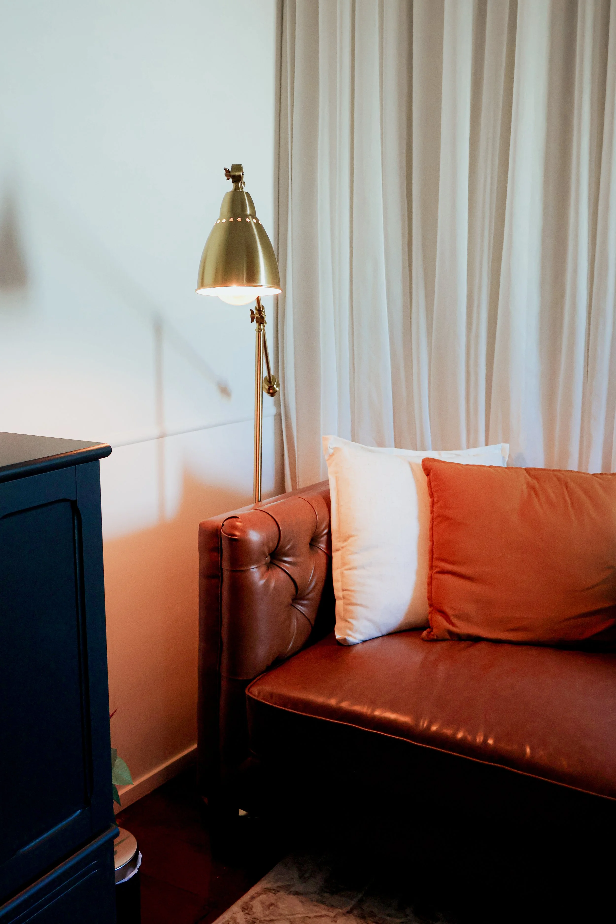 A corner of a cozy living room with a brown leather tufted sofa, white and rust-colored pillows, a brass floor lamp, and a cream curtain in the background.