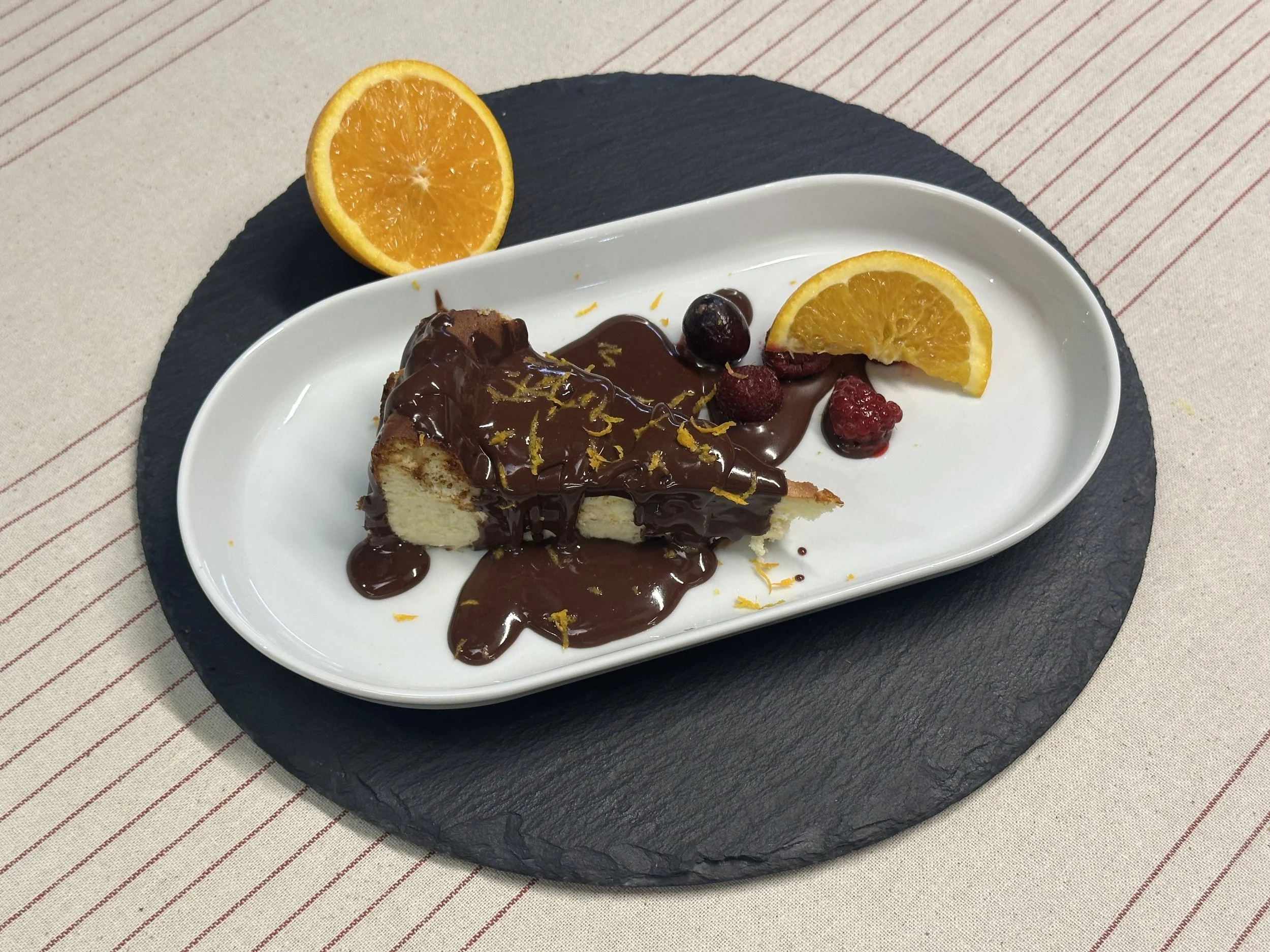 Slice of cheesecake with chocolate sauce, garnished with orange zest, served with orange wedge and berries on a white plate, placed on a black slate serving board.