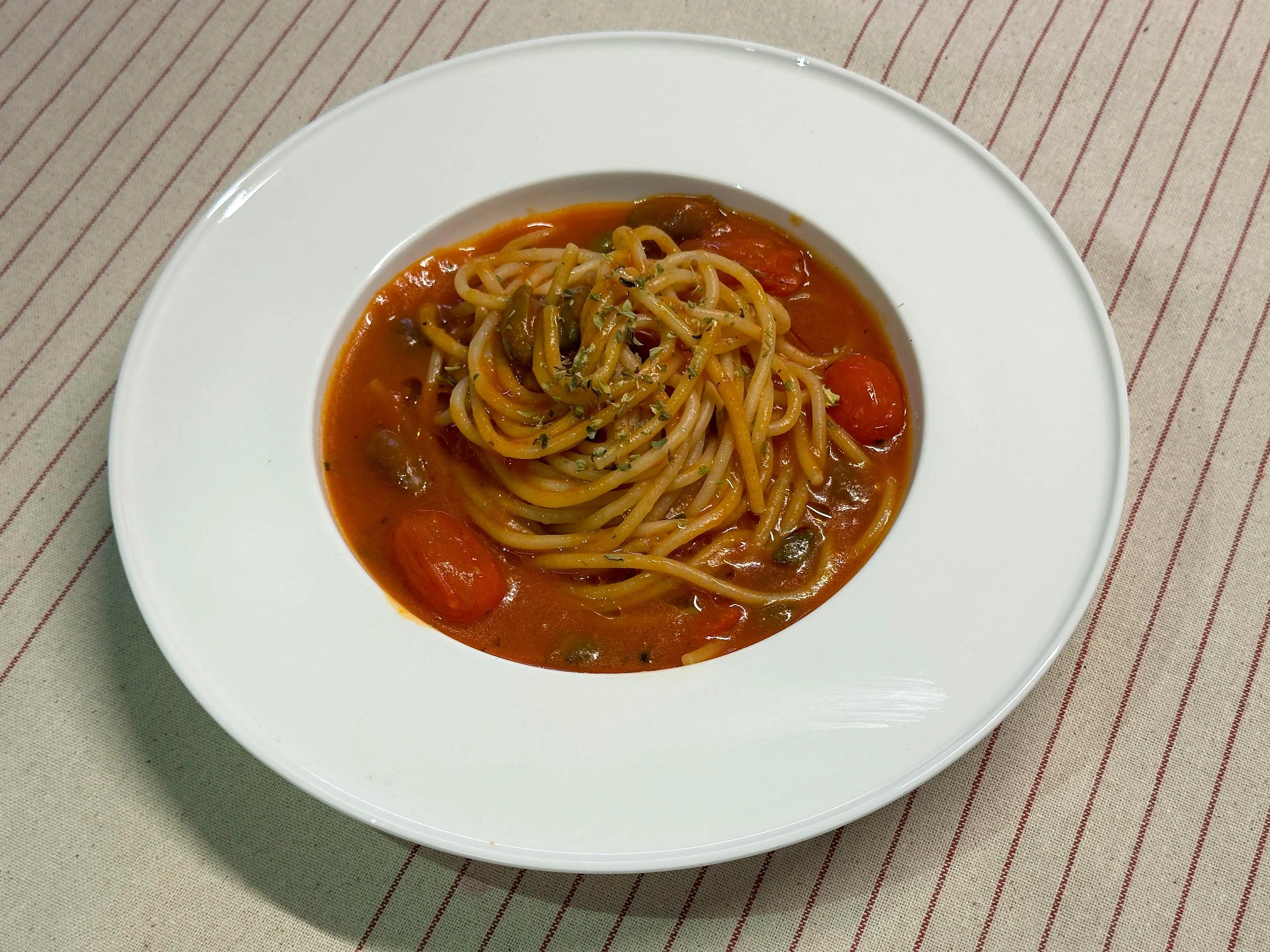 Plate of spaghetti with tomato sauce, cherry tomatoes, olives, and herbs on a striped tablecloth.
