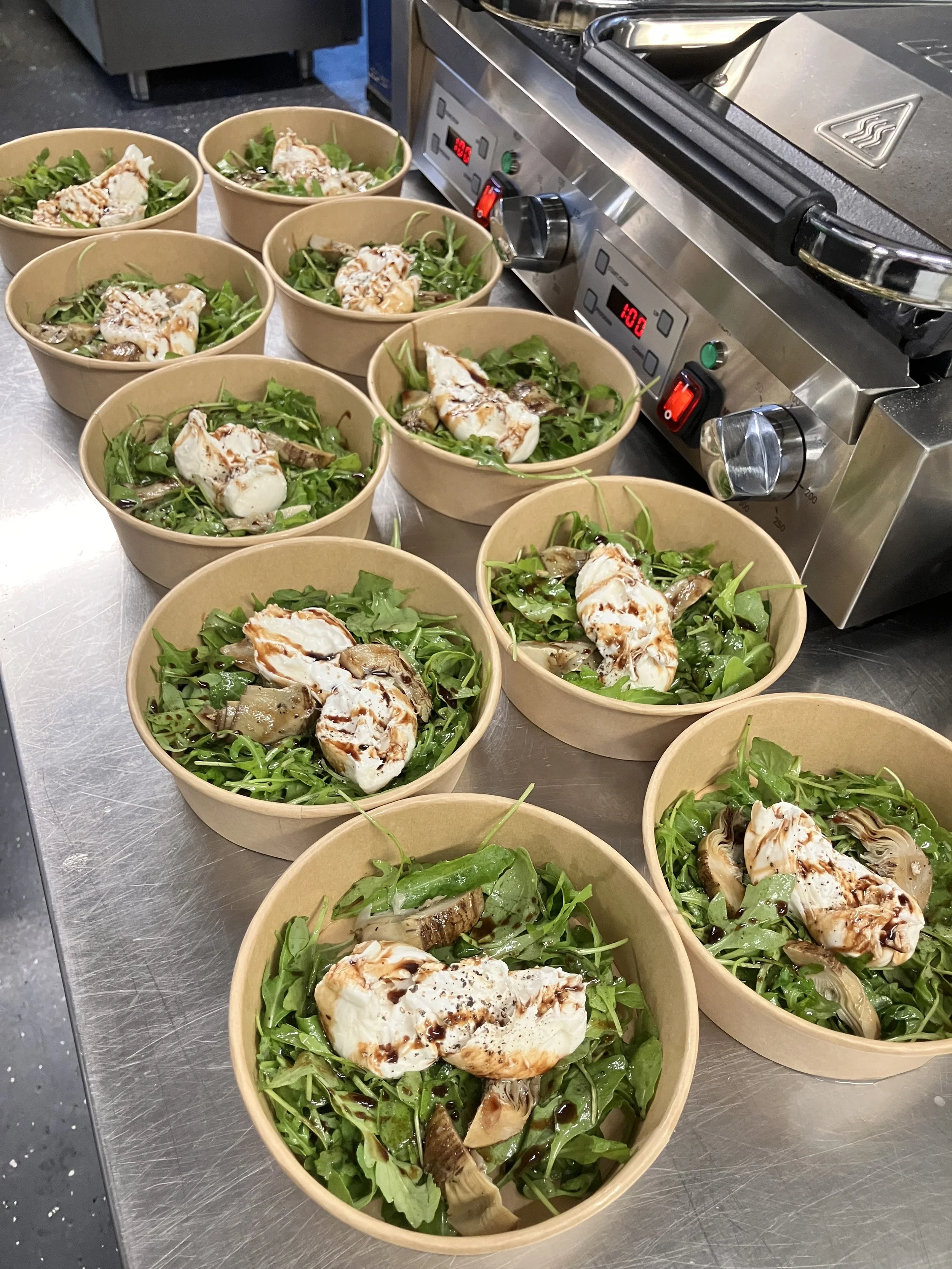 Meerdere zorgvuldig samengestelde salades met verse bladgroenten, geselecteerde kazen, champignons en een verfijnde balsamicoglazuur, gepresenteerd op een roestvrijstalen werkblad in een professionele keukenomgeving. Deze opstelling weerspiegelt effi