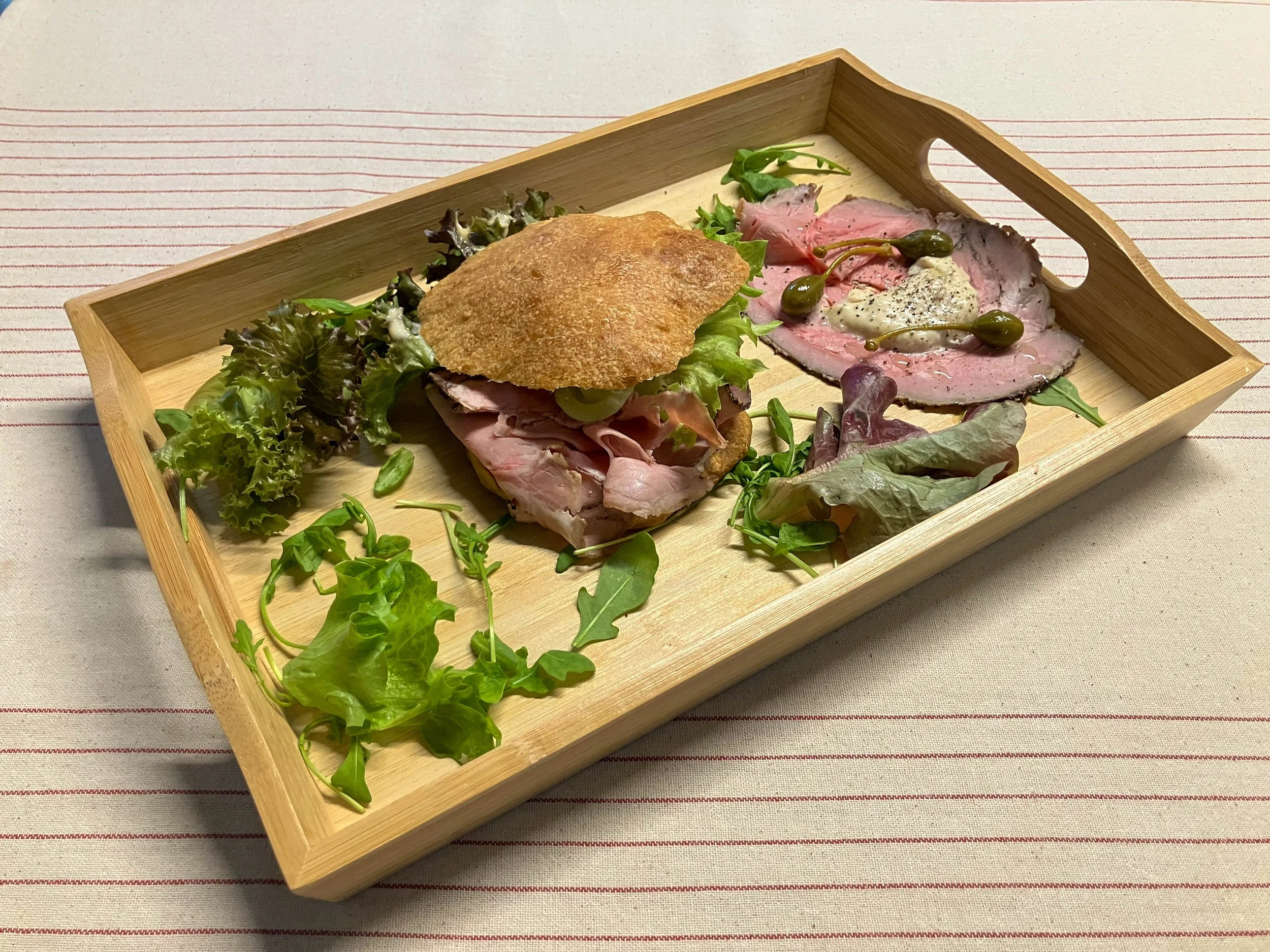 Een stijlvol gepresenteerde houten serveerplank met een rijk belegde sandwich van verse groenten, zorgvuldig geselecteerde vleeswaren en knapperig brood. Aangevuld met extra vleeswaren, greens en verfijnde toppings aan de zijkant, straalt deze presen