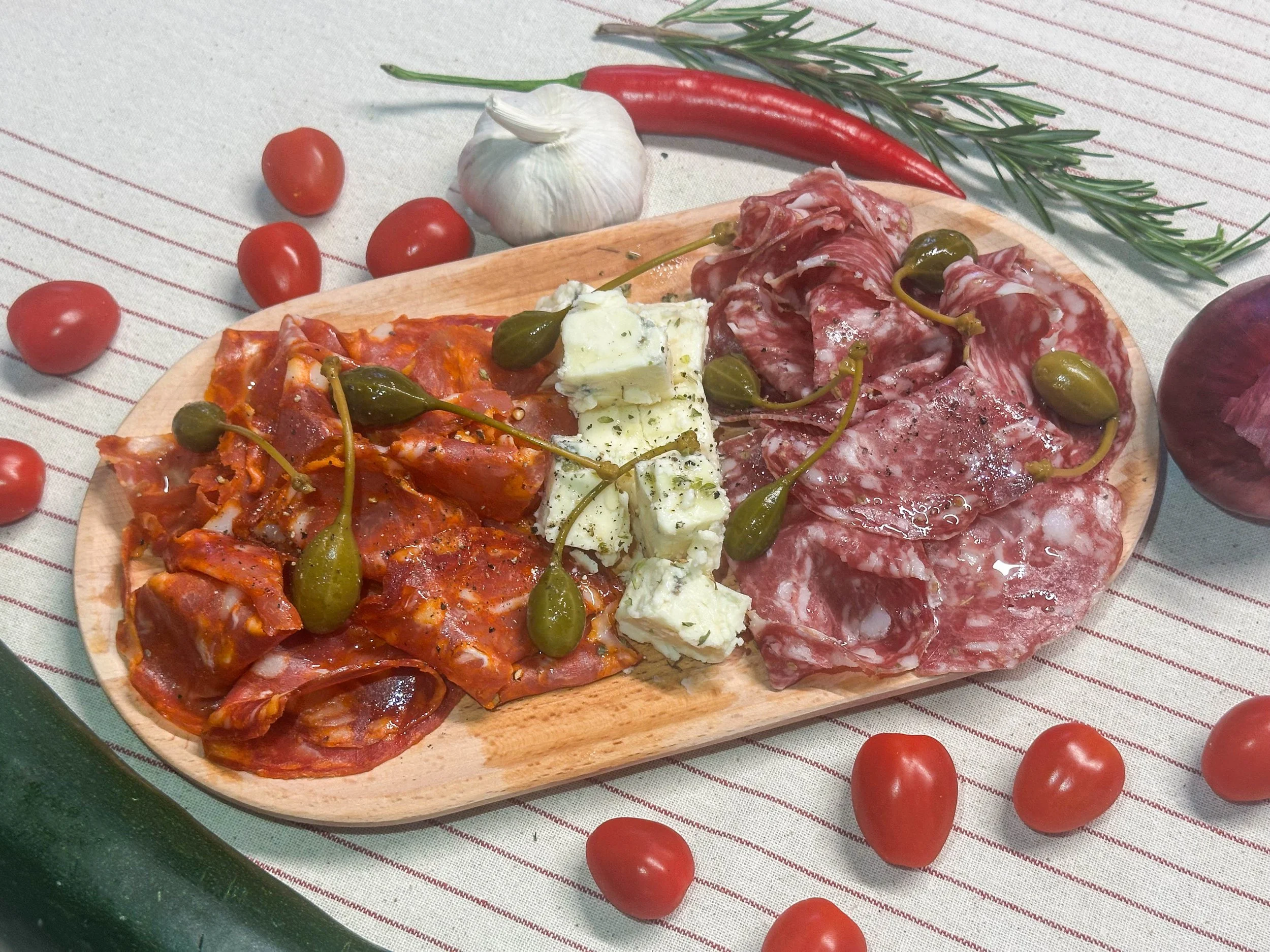 A wooden platter with assorted cured meats, blue cheese, and small green peppers, surrounded by cherry tomatoes, garlic, rosemary, red onion, and a cucumber.