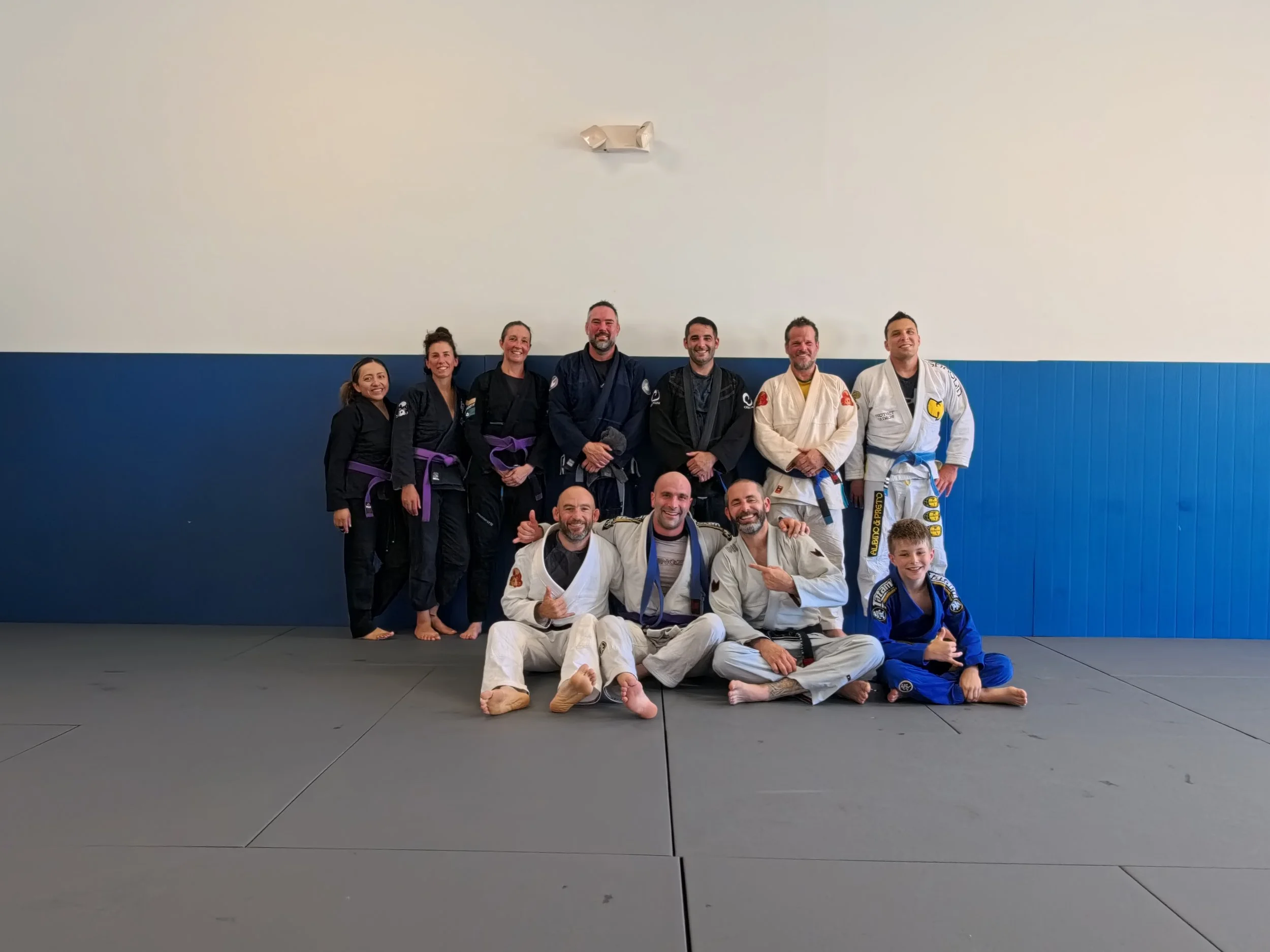 Group of martial artists, including children and adults, posing together in jiu-jitsu gis on a training mat in a martial arts dojo, smiling and making gestures.