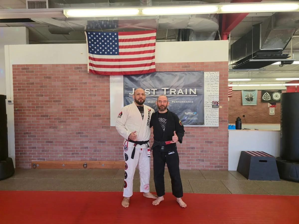 Two men in Brazilian Jiu-Jitsu gis standing on a mat in a gym, giving thumbs up. An American flag hangs on a brick wall behind them. The wall has a large banner that says "Just Train" and a whiteboard with writing on it.